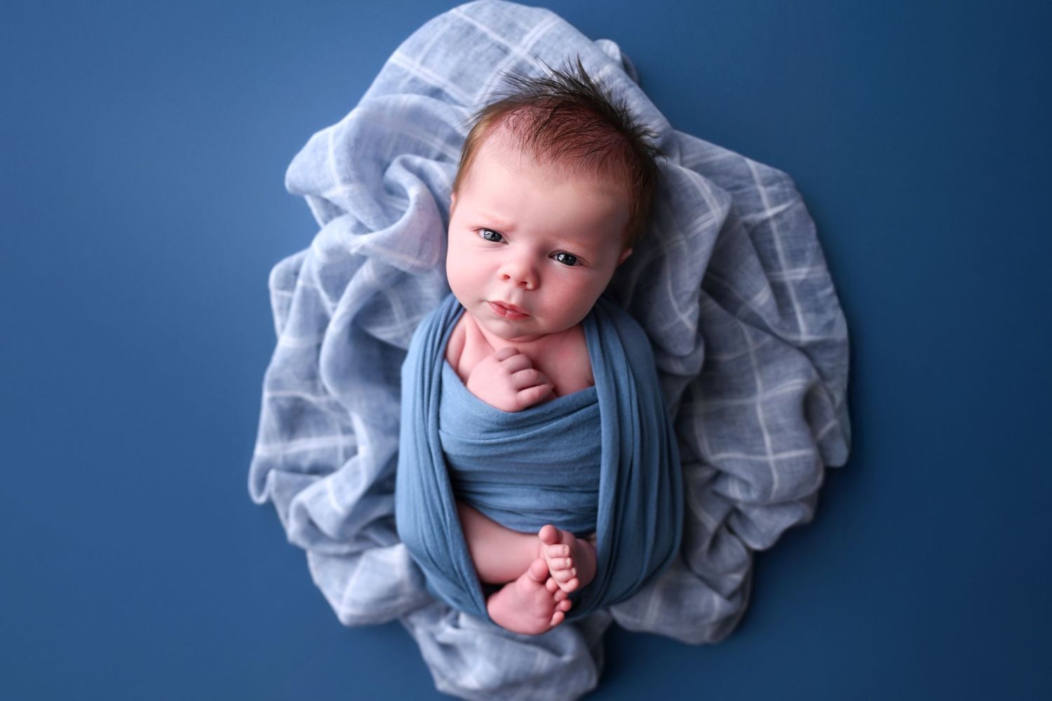 Baby wrapped up over a soft plaid blanket, looking directly into the camera with a pensive face