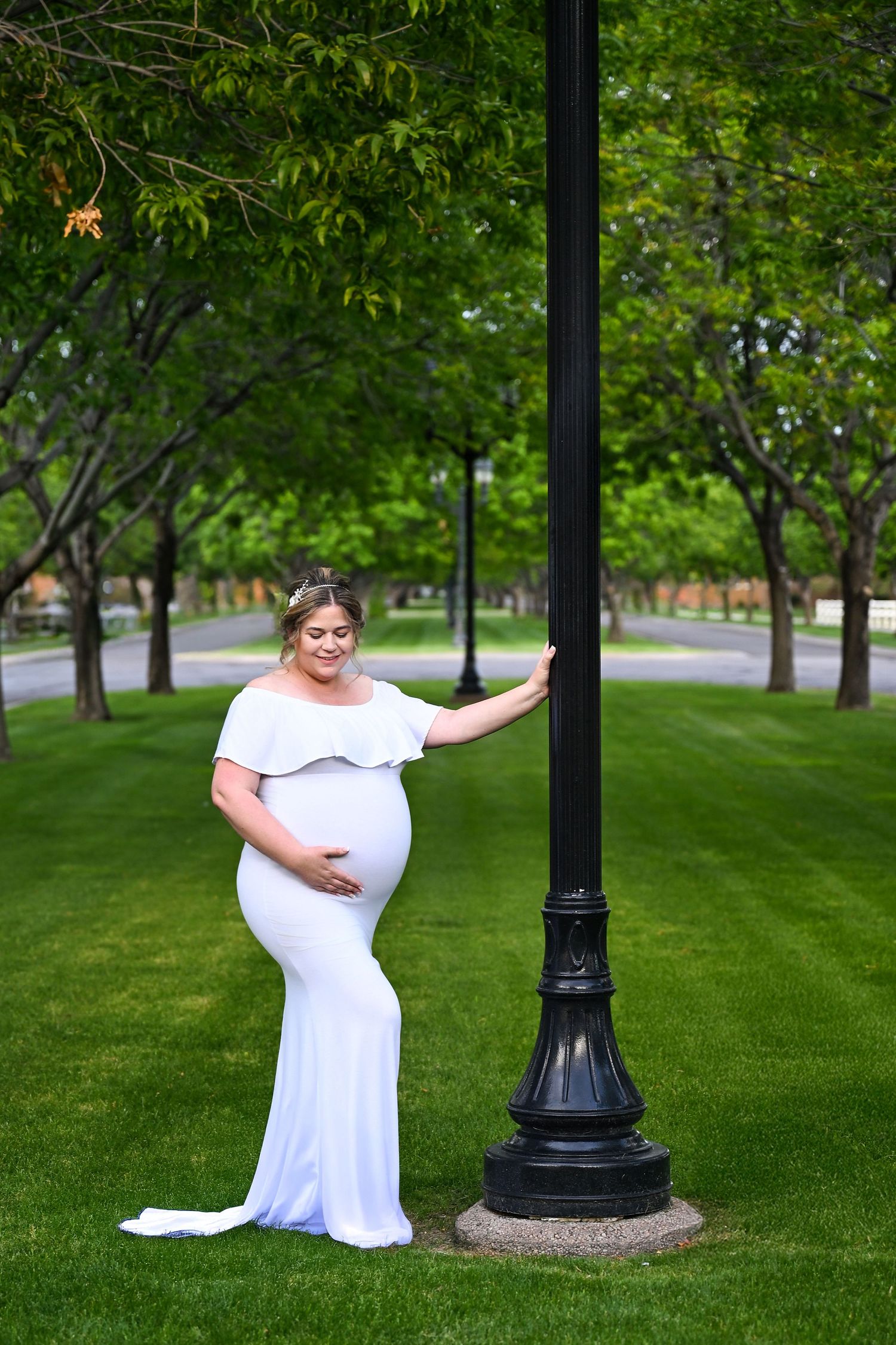 A woman in a long white gown standing holding onto a Gilbert Arizona Light post while looking down at her pregnant belly