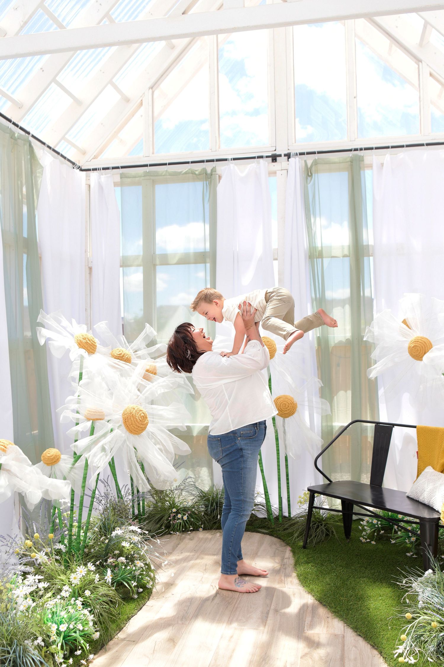Mother Holds toddler up above her head in white greenhouse laughing surrounded by flowers on an Arizona spring morning sunny day