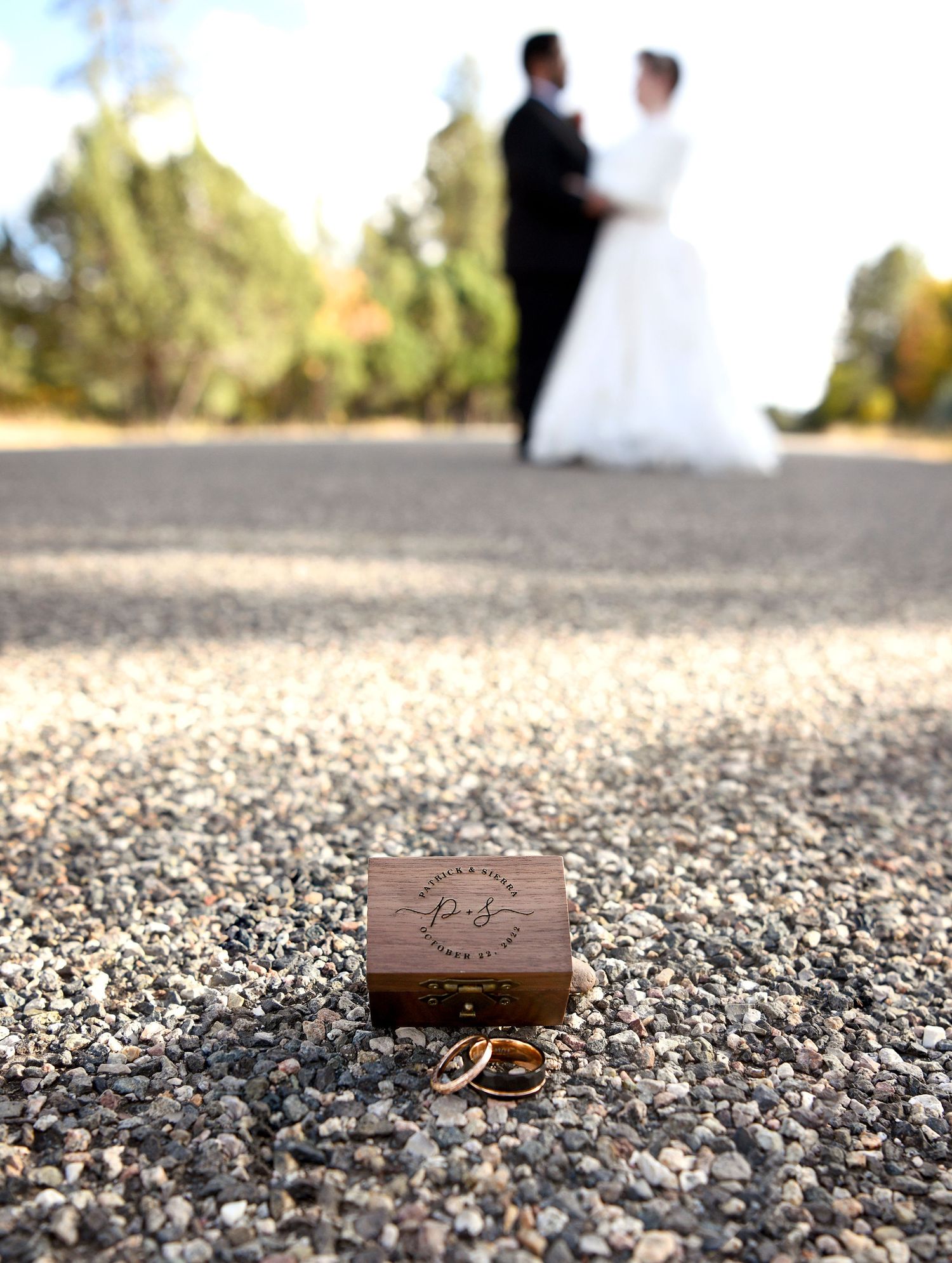 Detail shot of wedding rings and wooden box on the floor at golden hour with bride and groom in the background.