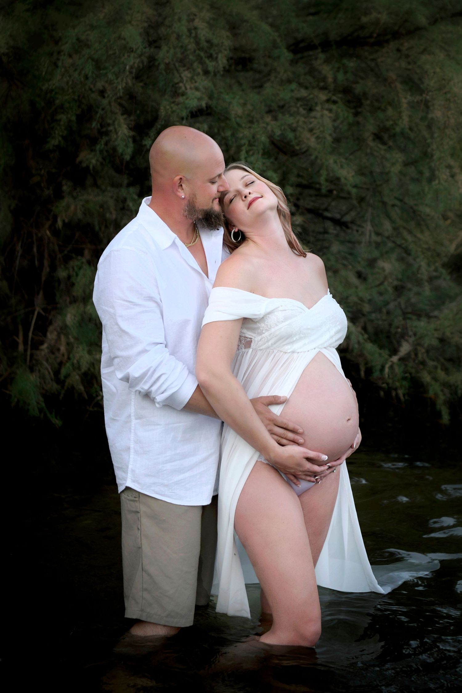 Couple stands  in the Salt River enjoying the moment wearing matching white outfits. Her pregnant belly is showing
