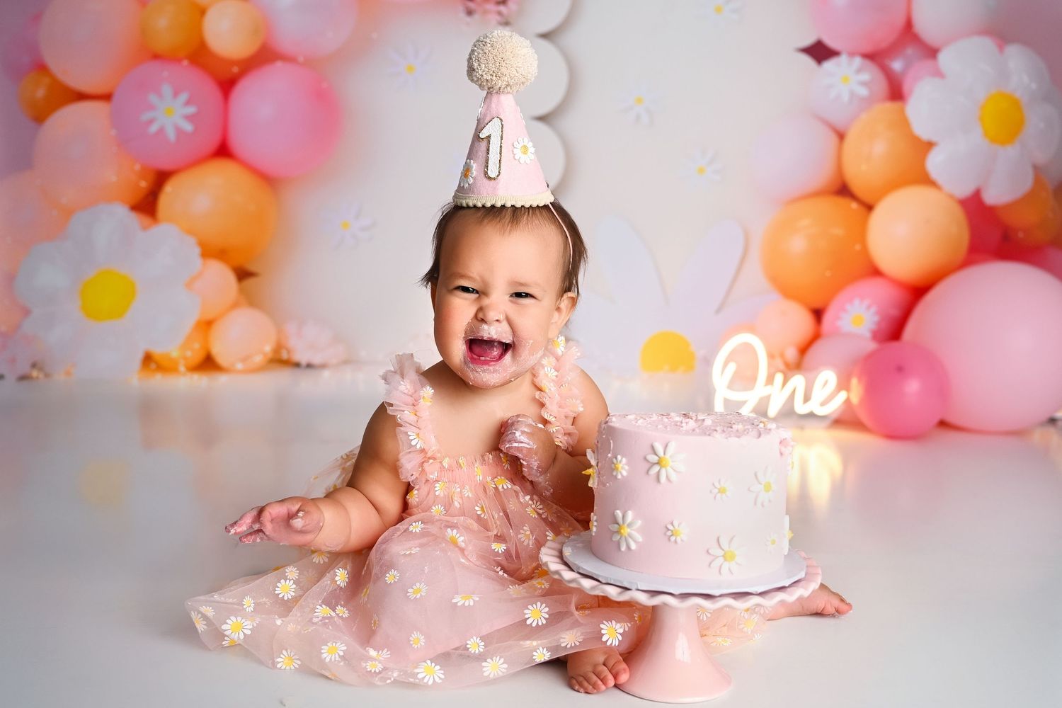 Laughing one year old baby girl with pink daisy cake and balloon garland for Cake Smash Photos