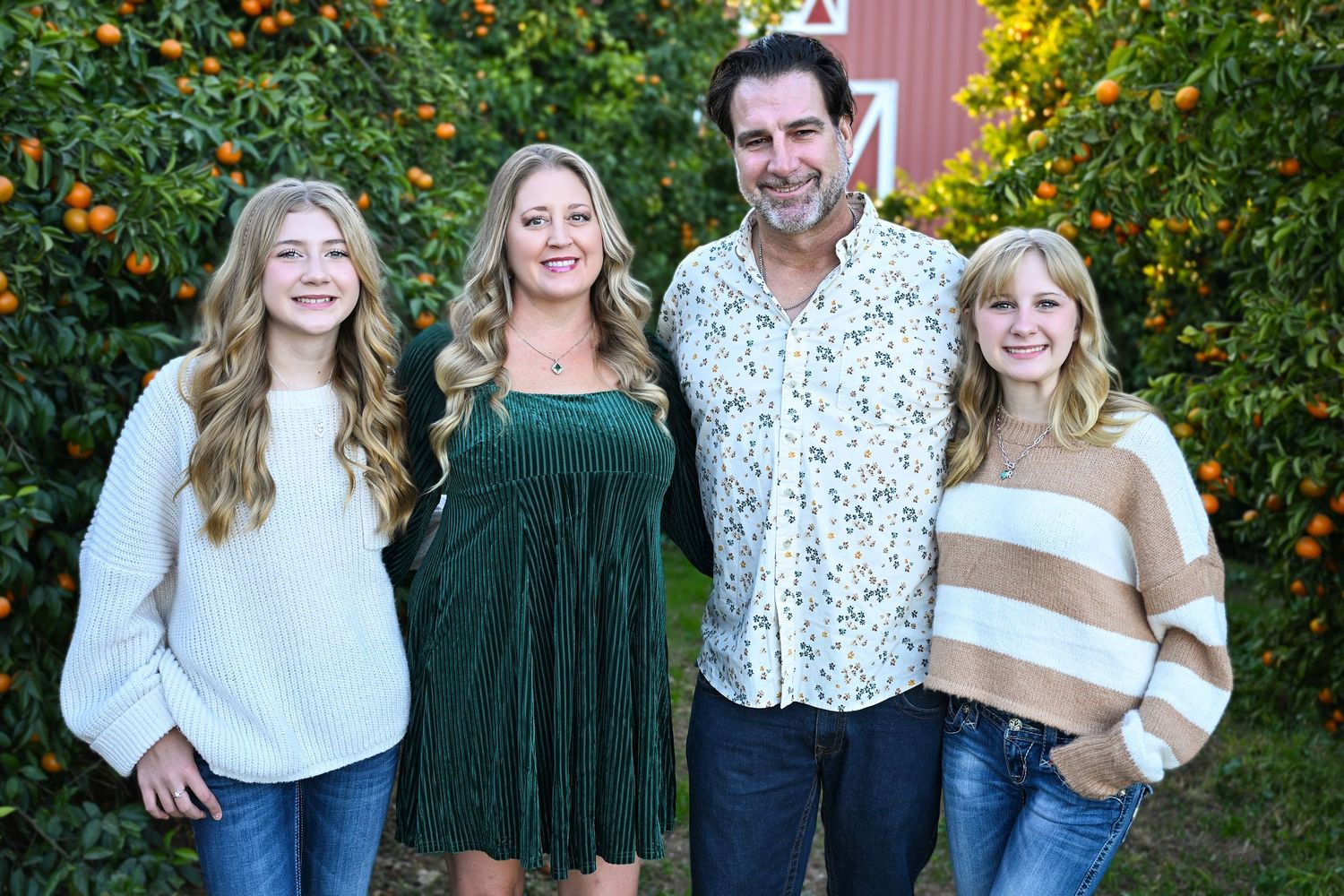 Family of four stand in Queen Creek Citrus grove with teenage daughters in front of big red barn