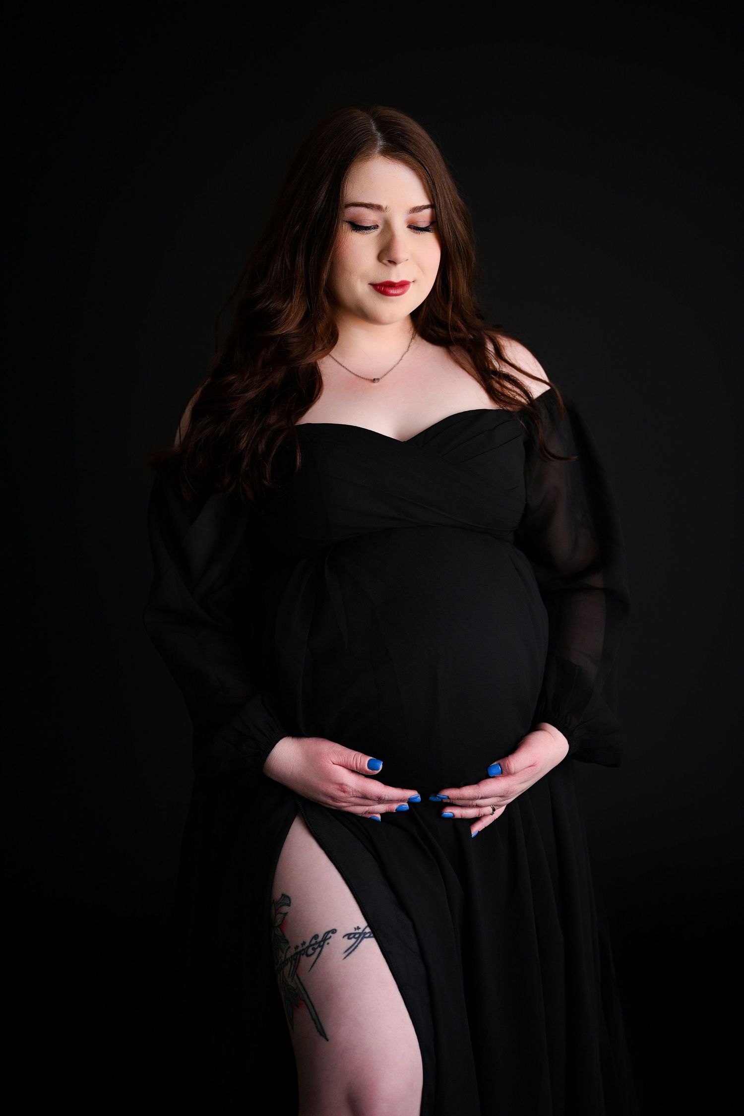 Woman in black staring down at her pregnant belly in an all black photography studio with a large tattoo on her leg
