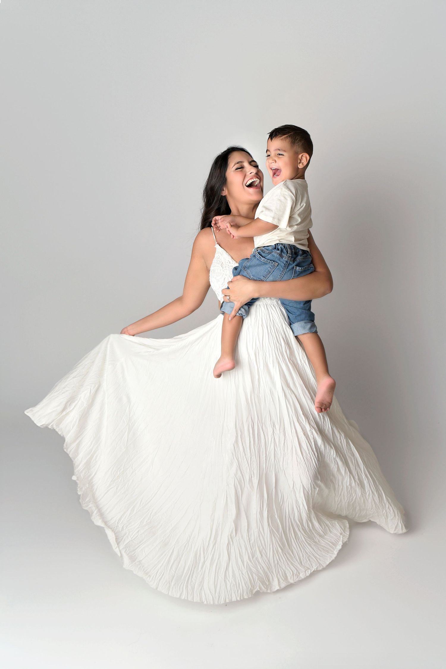 Mother and Son dancing around in white gown on white seamless paper in Phoenix photography studio for maternity session.