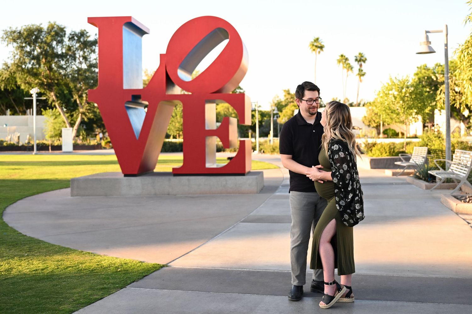 Couple stands laughing together in front of iconic Scottsdale LOVE sign. The woman is pregnant and wearing a gown for her Maternity pics