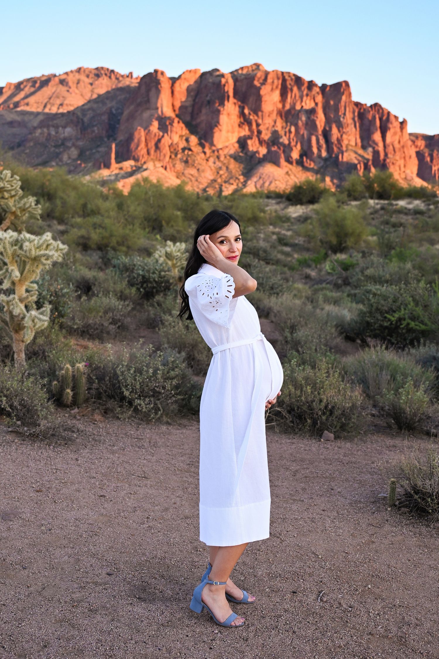 Mother brushes hair away from face while standing in the Arizona desert. Golden Hour at the Superstition Mountains in Apache Junction, AZ.