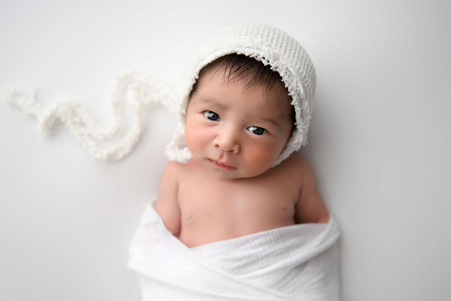 All white image of newborn baby boy looking into the camera wearing a white bonnet