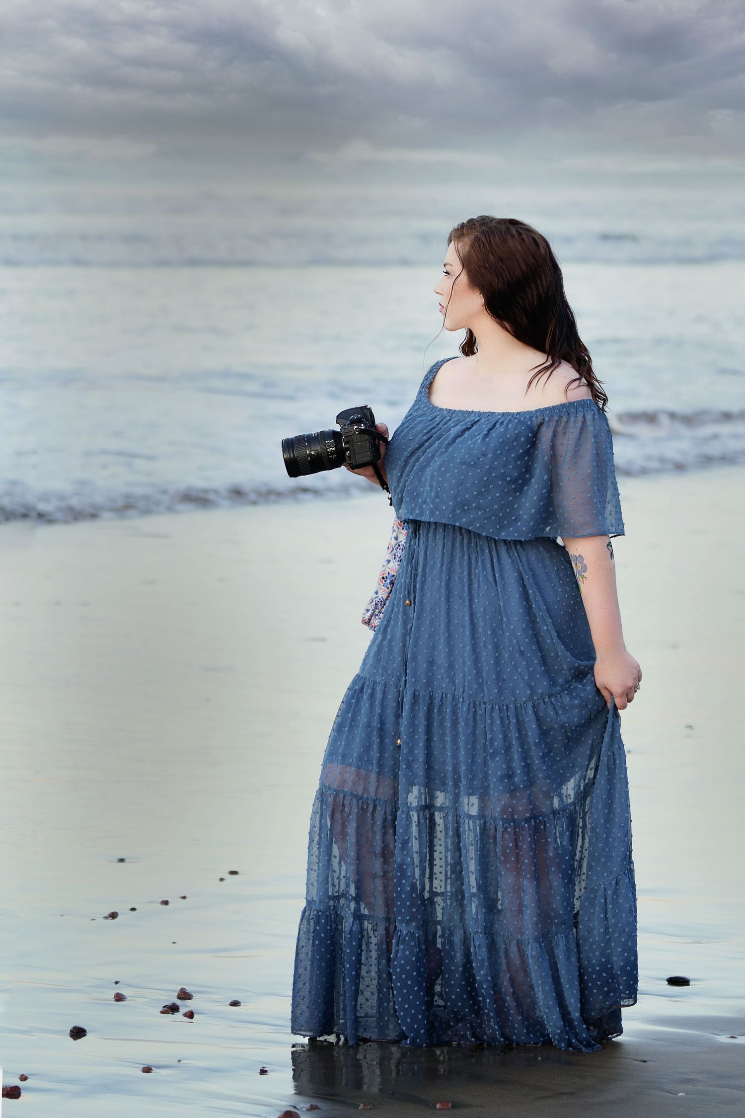 Photographer stands holding camera in blue gown looking out over the water in moody portrait