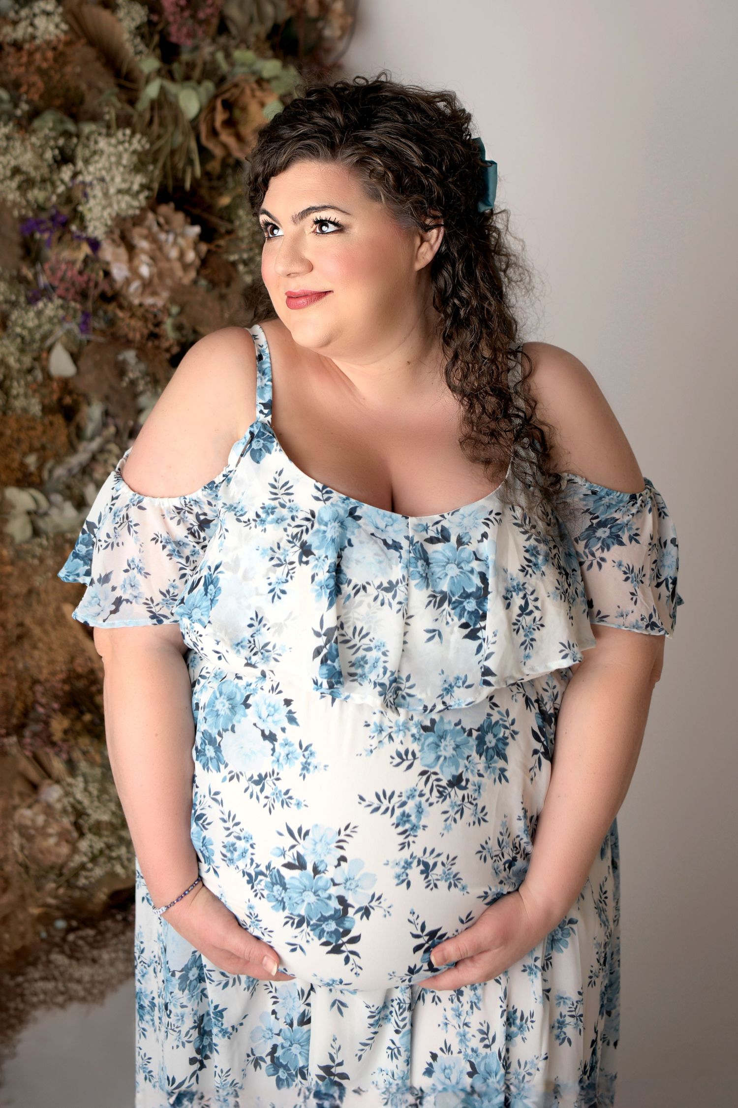 Beautiful curly haired woman stands holding her pregnant belly with a flower wall behind her. She looks off camera smiling to someone unseen