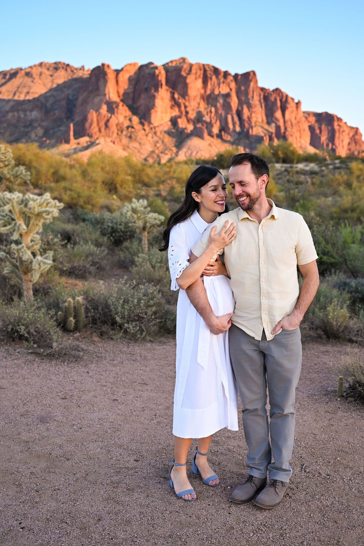 Husband and wife smiling and touching her pregnant belly in a desert at sunset.