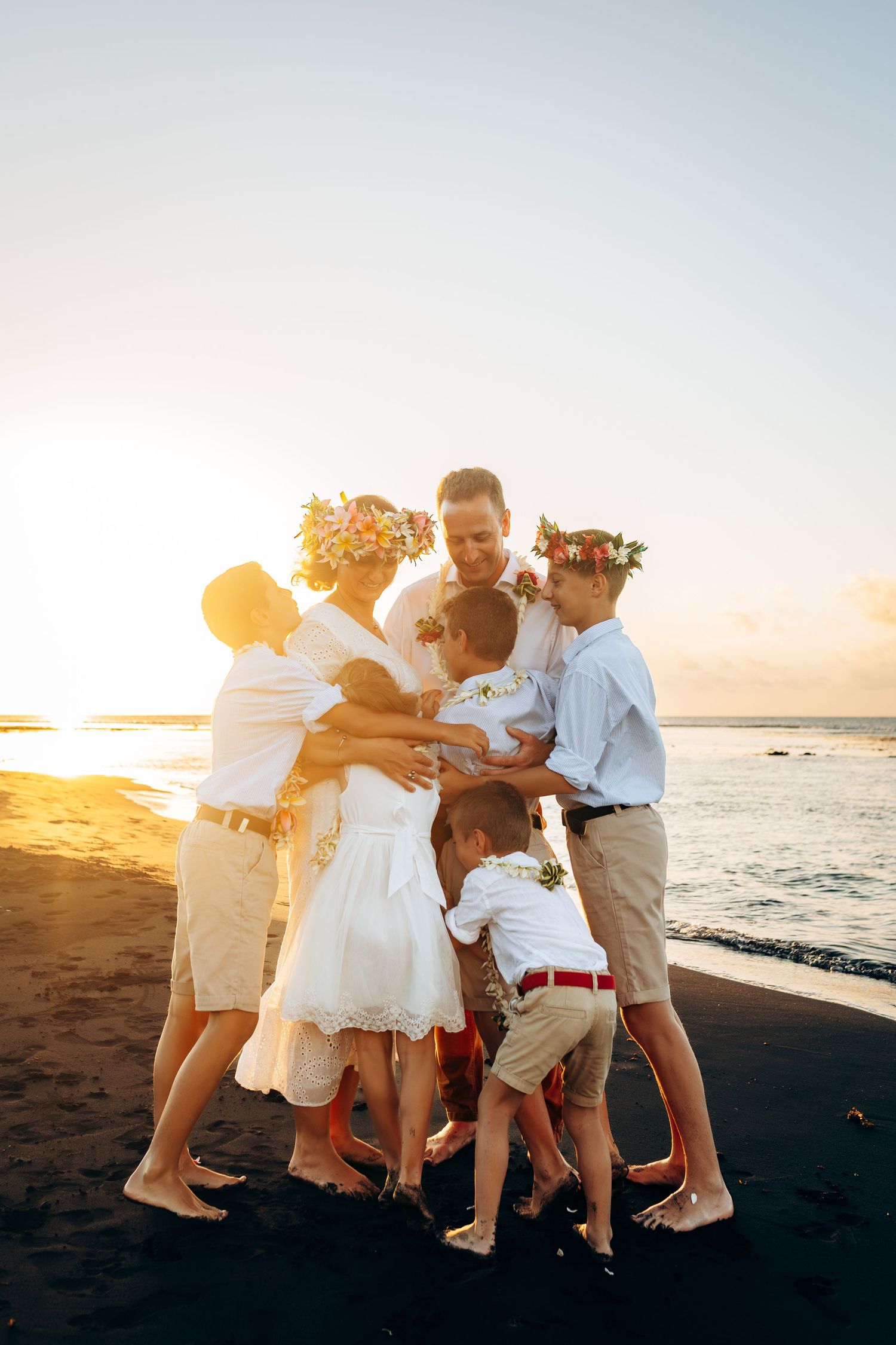 Famille en tenues blanches avec des couronnes florales s'embrassant sur une plage de sable noir au coucher de soleil doré.