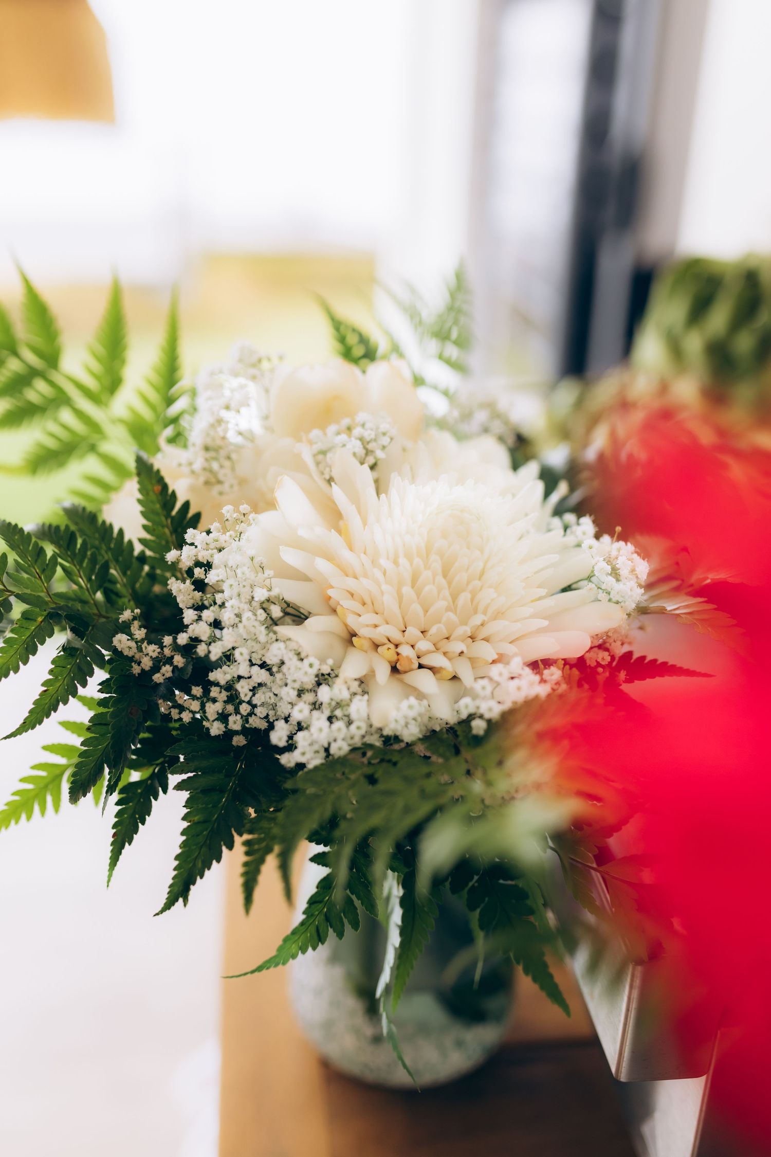 Elegant white chrysanthemum and baby's breath bouquet with green ferns in glass vase by window.