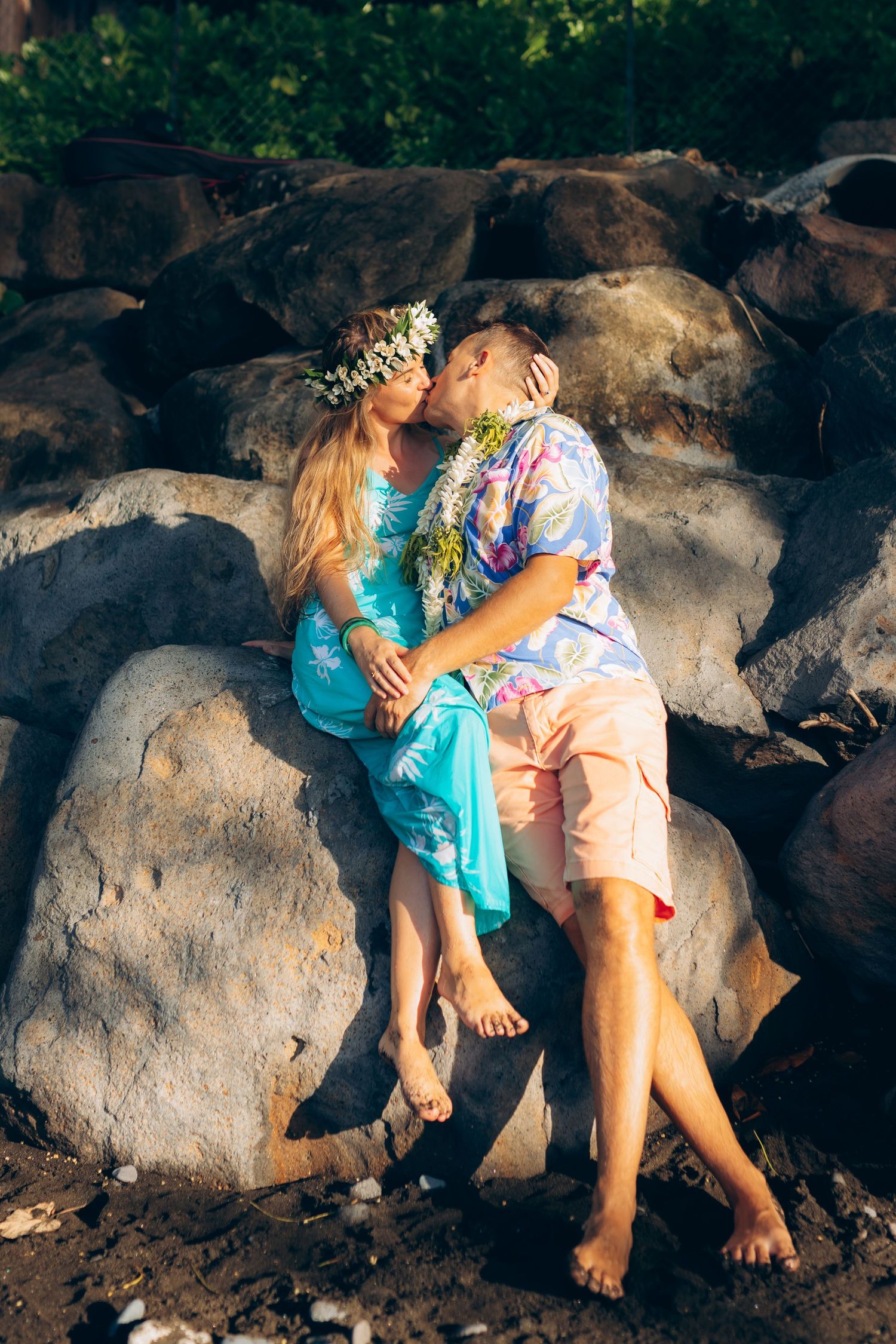 Couple partageant un baiser assis sur des rochers dans un jardin tropical au coucher du soleil.