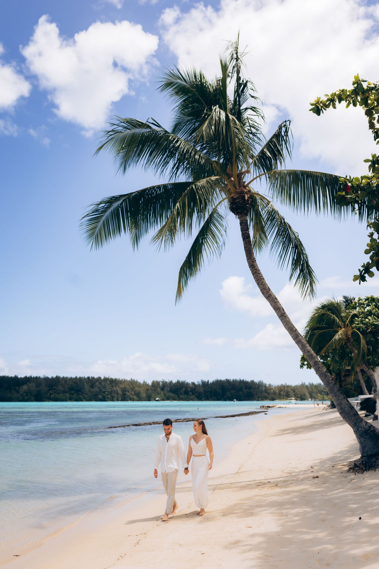 Couple de mariage en blanc se tenant la main marchant sur une plage tropicale sous un palmier.