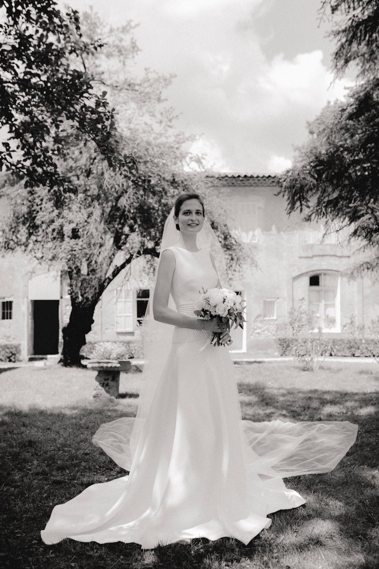 Bride in elegant gown with veil holding bouquet stands in garden of historic venue.