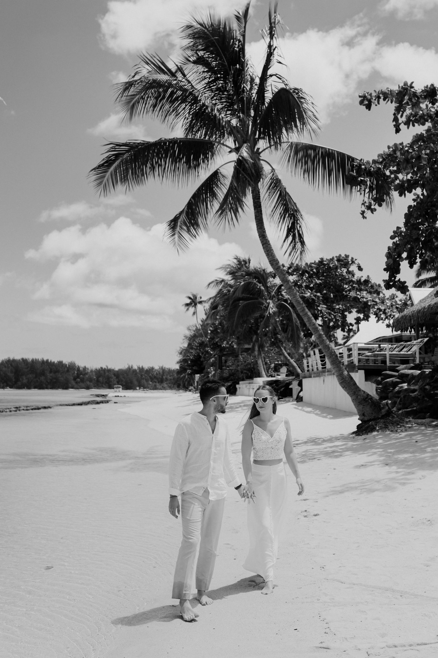 Photo en noir et blanc d'un couple en blanc marchant sur une plage tropicale avec des palmiers.