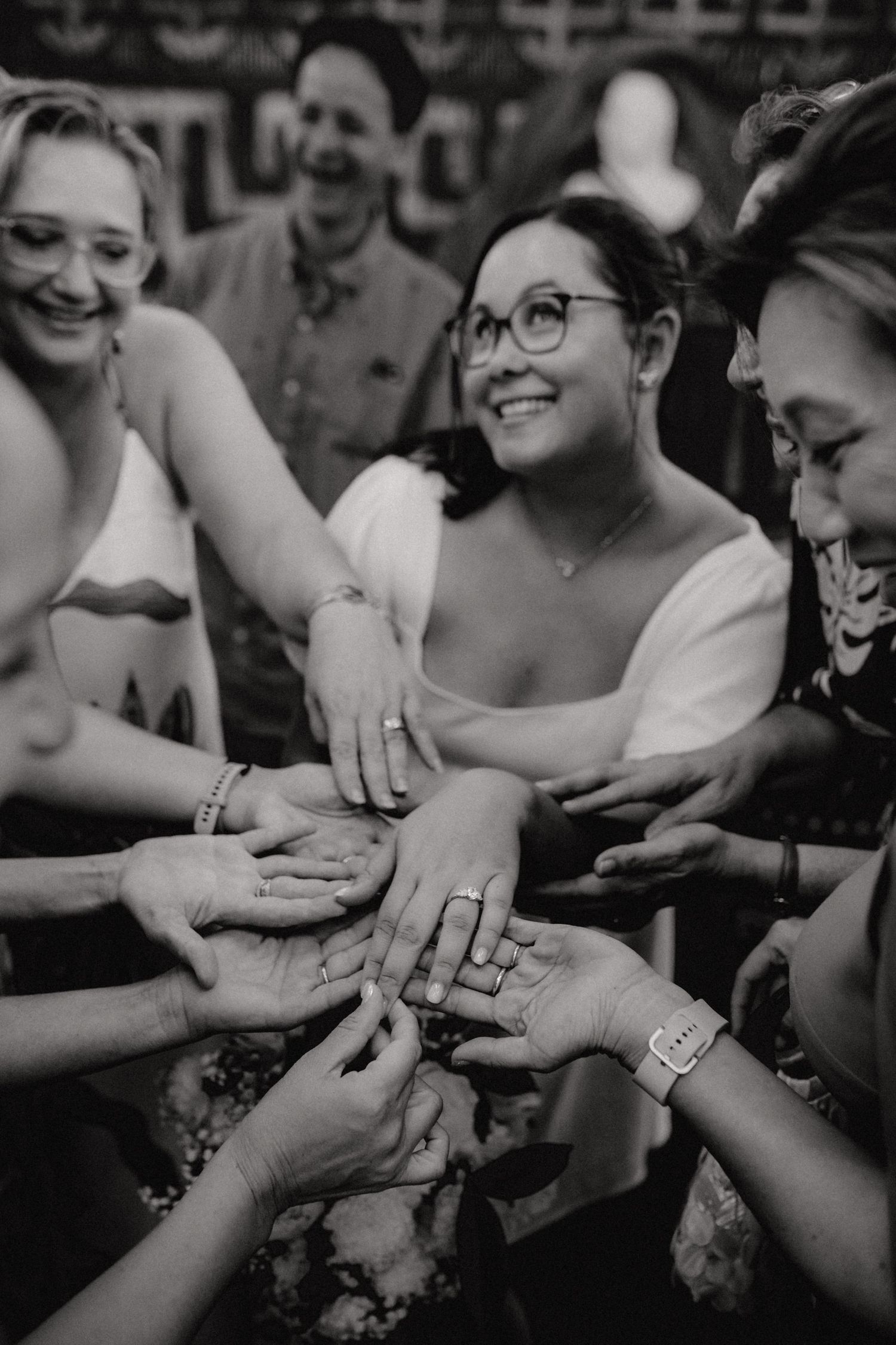 Black and white photo of smiling wedding guests showing off rings and jewelry at a celebration.