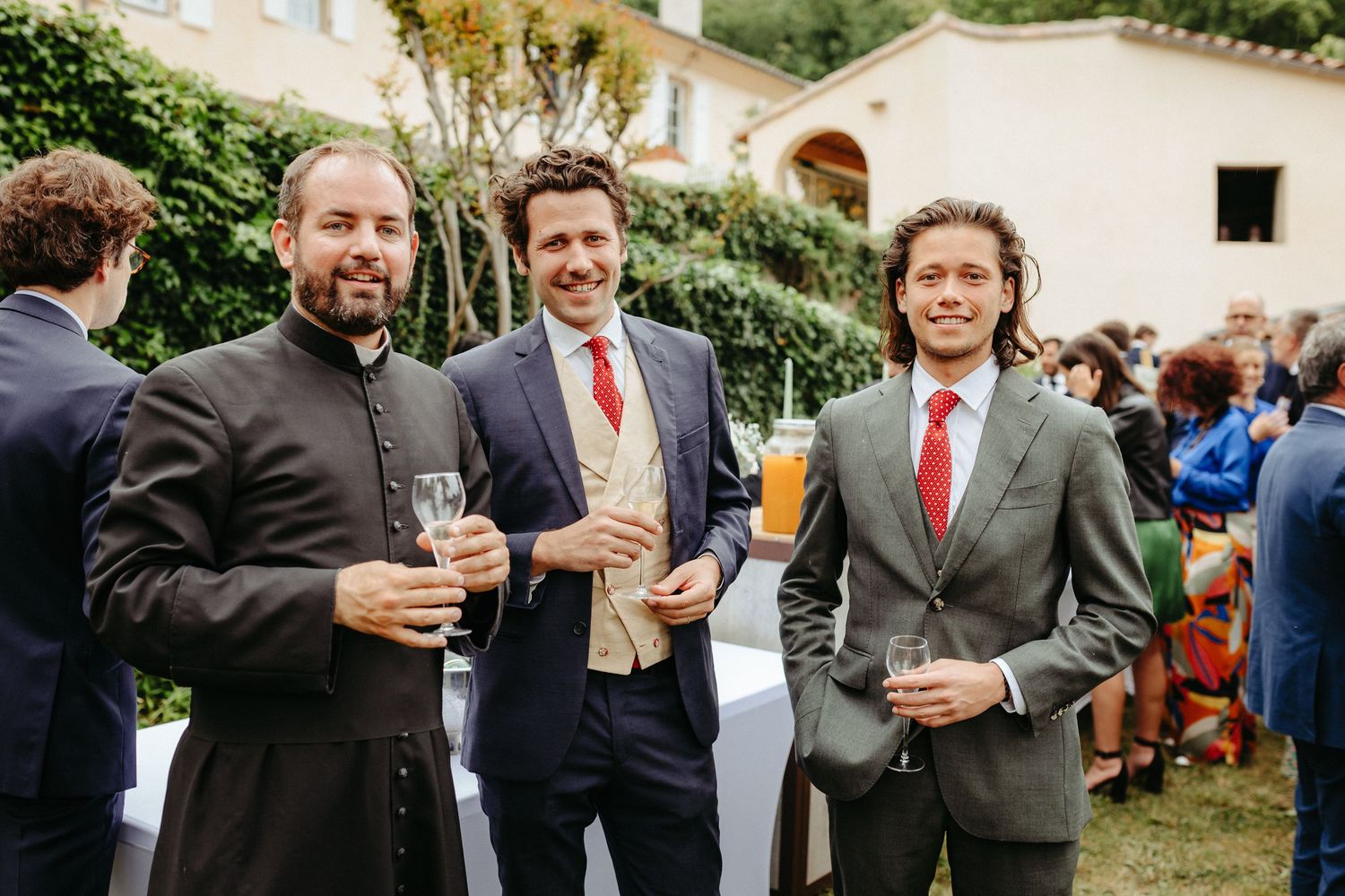 Trois hommes bien habillés en costume tenant des coupes de champagne lors d'un cocktail de mariage en plein air par une journée ensoleillée.