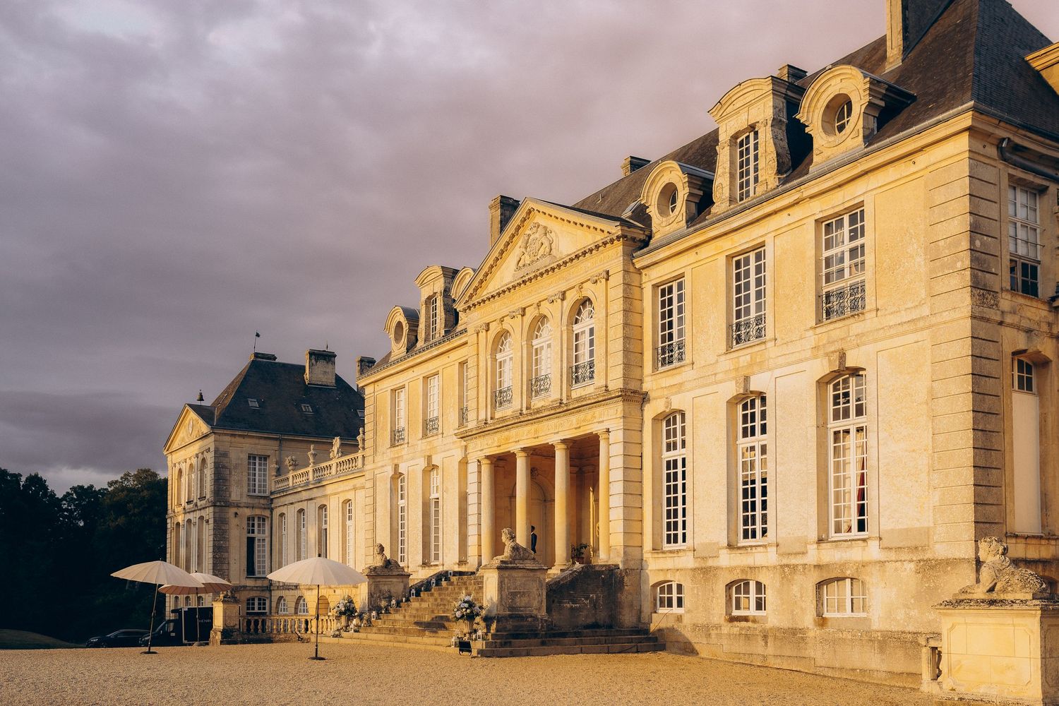 Grand château français avec des colonnes classiques et une façade en pierre ornée qui brille d'or sous un ciel nuageux spectaculaire.