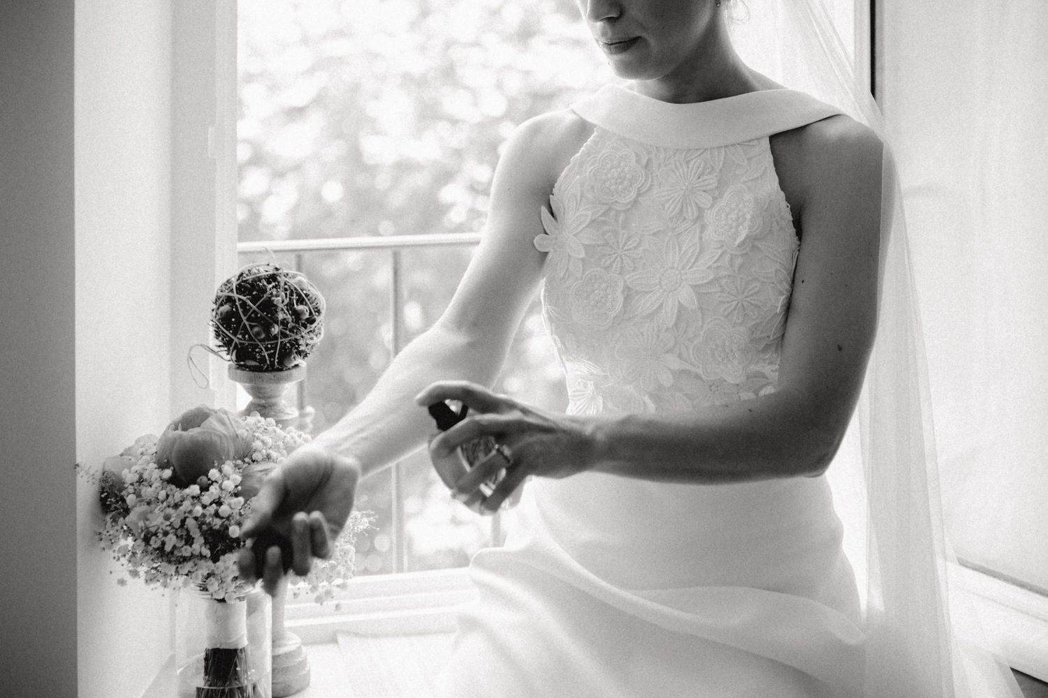 Photo en noir et blanc d'une mariée en robe en dentelle blanche tenant un bouquet près d'une fenêtre le jour de son mariage.
