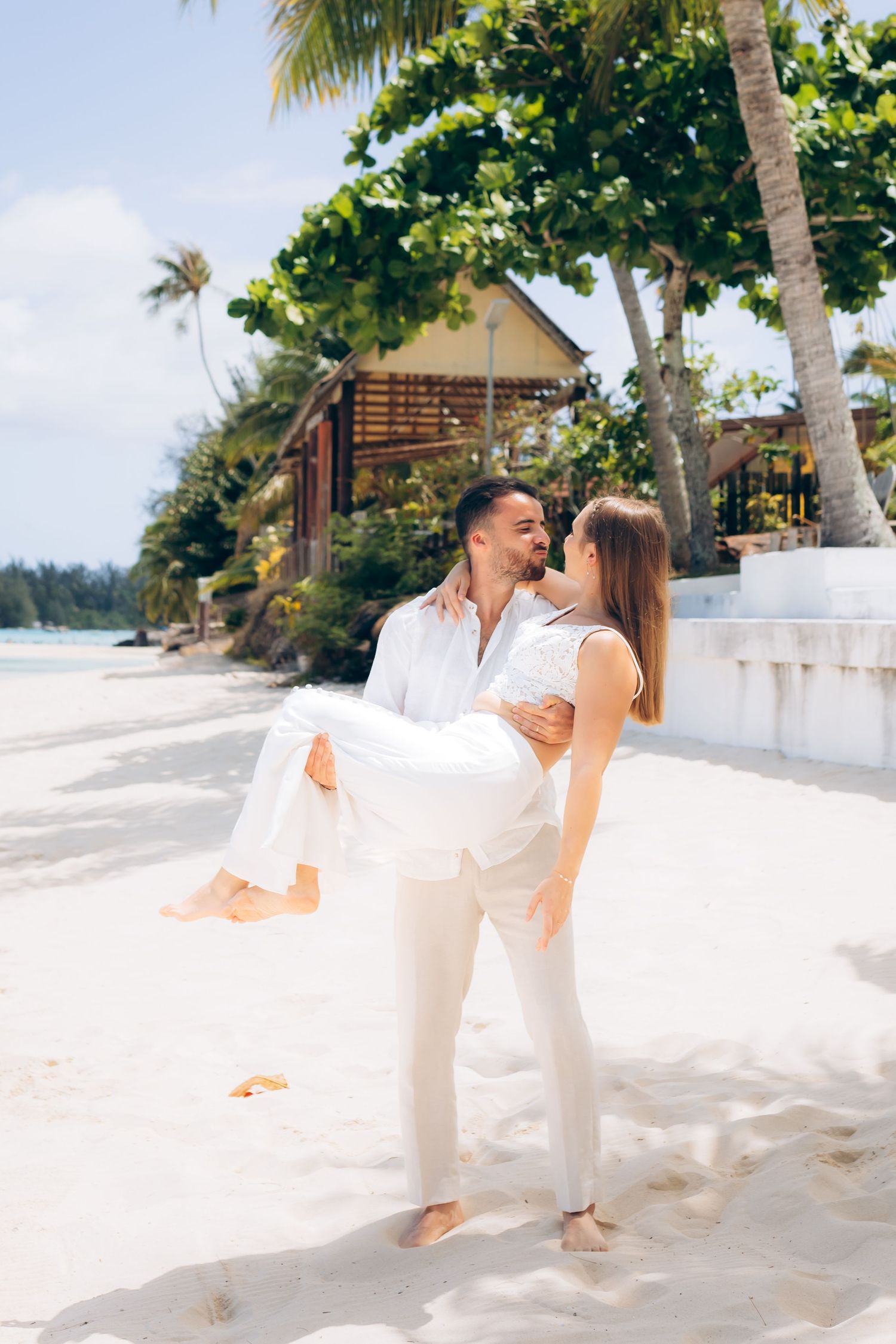 Marié portant une mariée en robe blanche sur une plage de sable blanc tropical avec des palmiers.