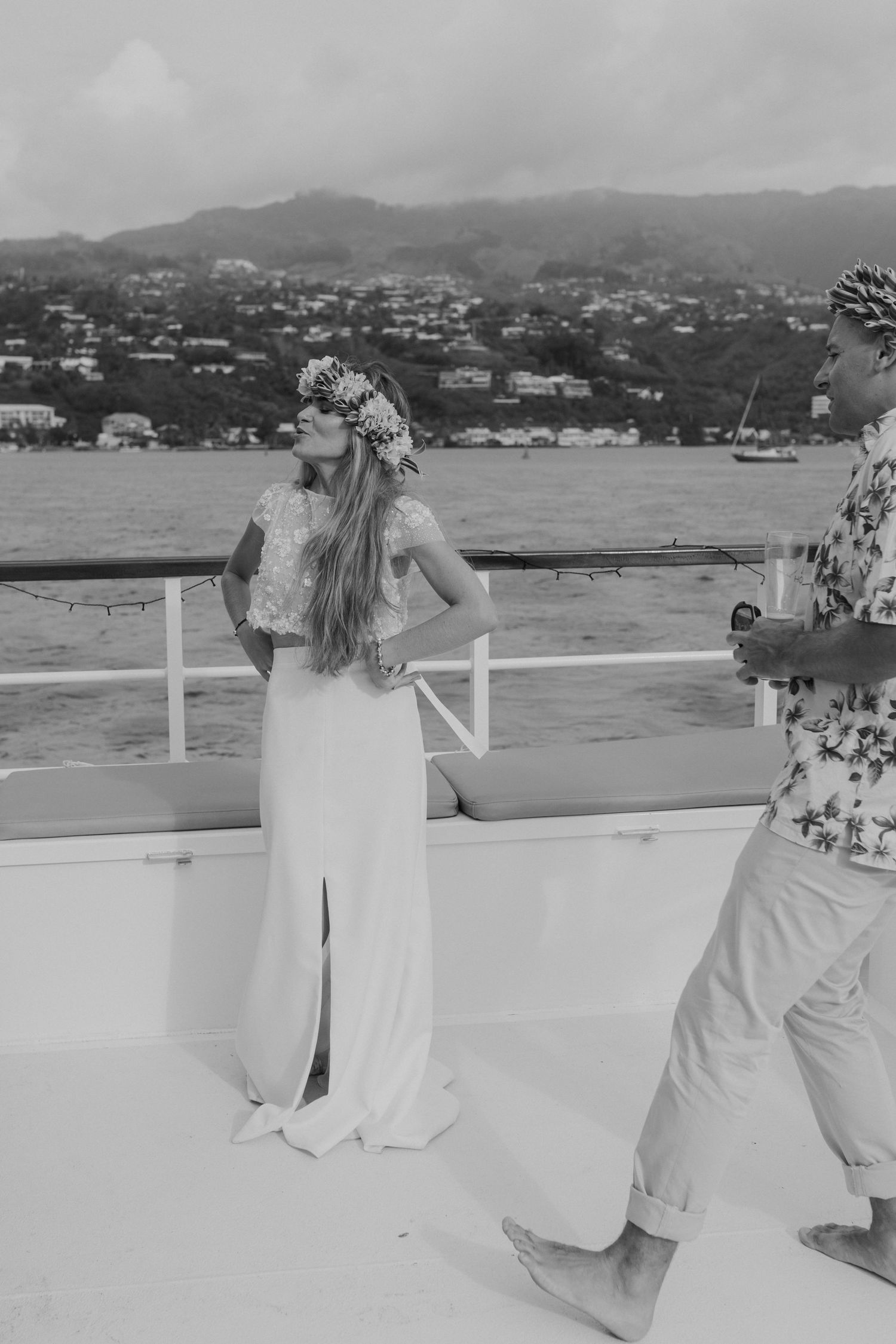 La mariée en robe blanche et couronne florale se tient sur le pont du yacht avec une toile de fond montagneuse.
