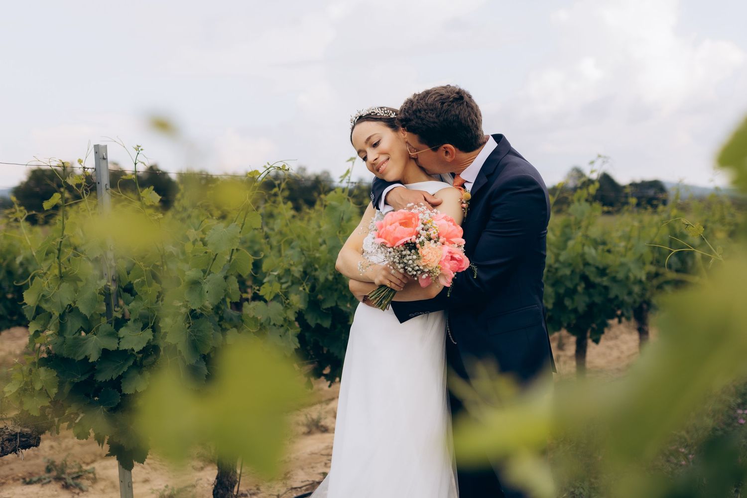 La mariée et le marié s'embrassent dans un vignoble, la mariée tenant un bouquet rose, le marié en costume bleu, douceur et poésie.