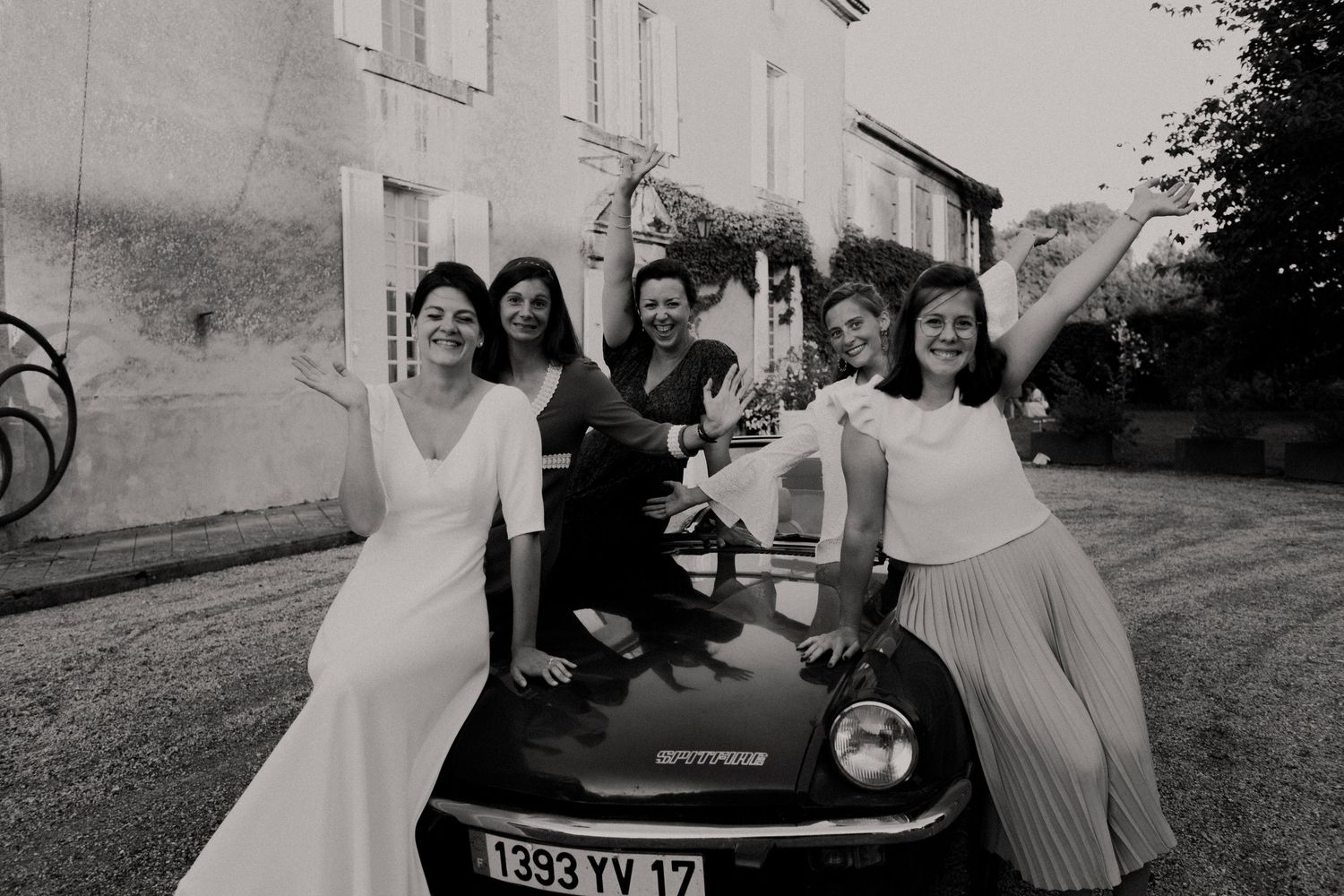 Photo en noir et blanc de la mariée et des demoiselles d'honneur en robes blanches posant joyeusement avec une voiture décapotable vintage.