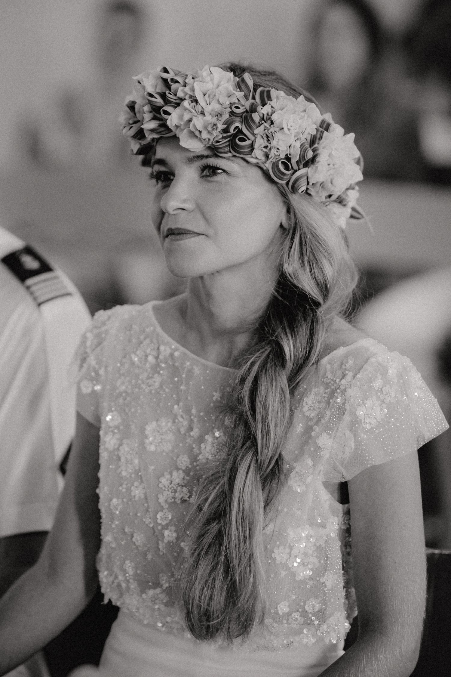 Bride wearing floral crown and sequined dress with braided hair at wedding ceremony in black and white.