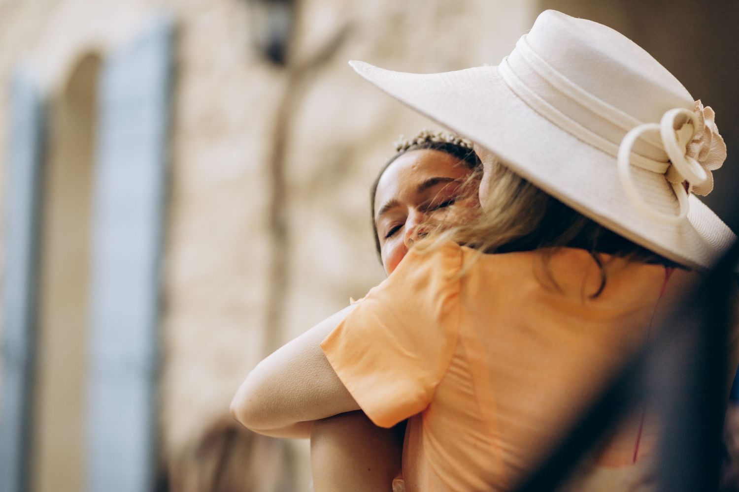 Deux femmes partagent une étreinte chaleureuse à l'extérieur, l'une portant un chapeau blanc et un accessoire floral avec une élégante robe