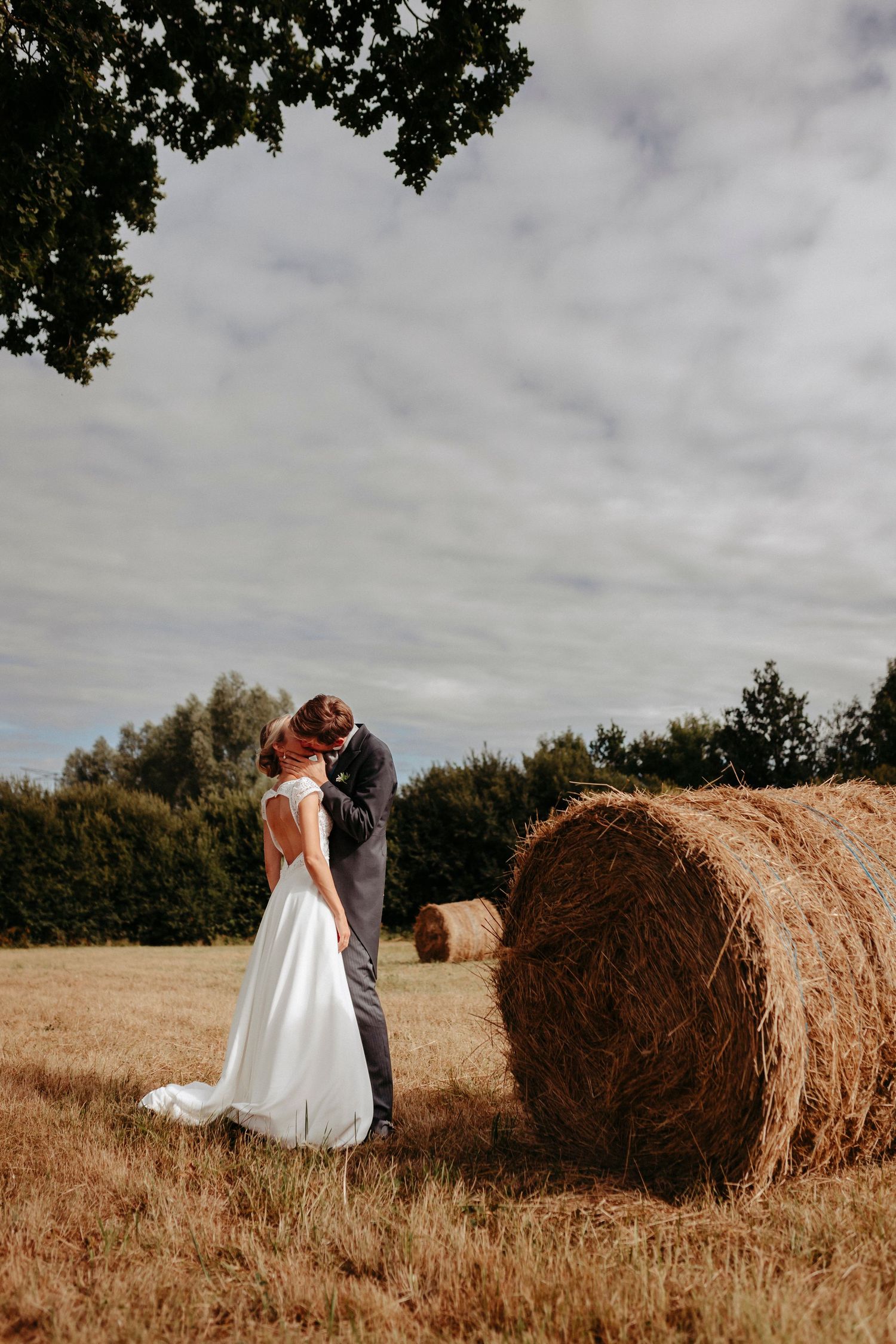 La mariée et le marié s'embrassent dans un champ doré à côté d'une balle de foin, avec des arbres et une toile de fond de ciel nuageux.
