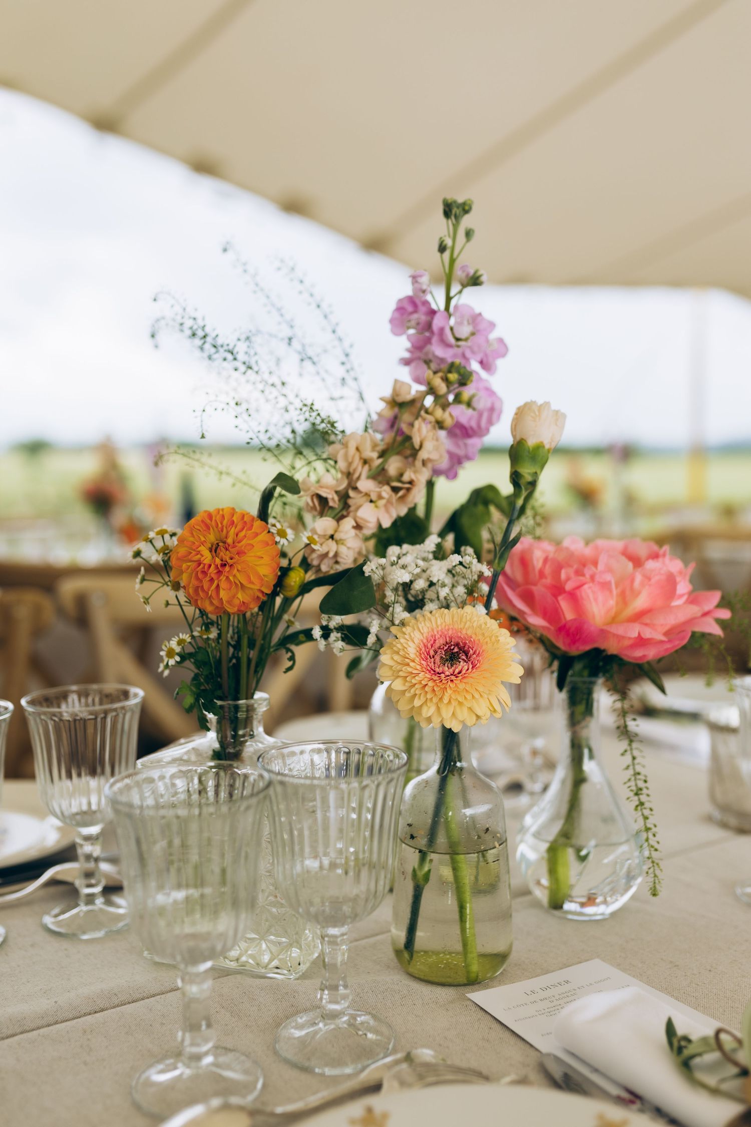 Centre de table de mariage coloré avec des dahlias orange, des pivoines roses et des fleurs sauvages dans des bouteilles en verre