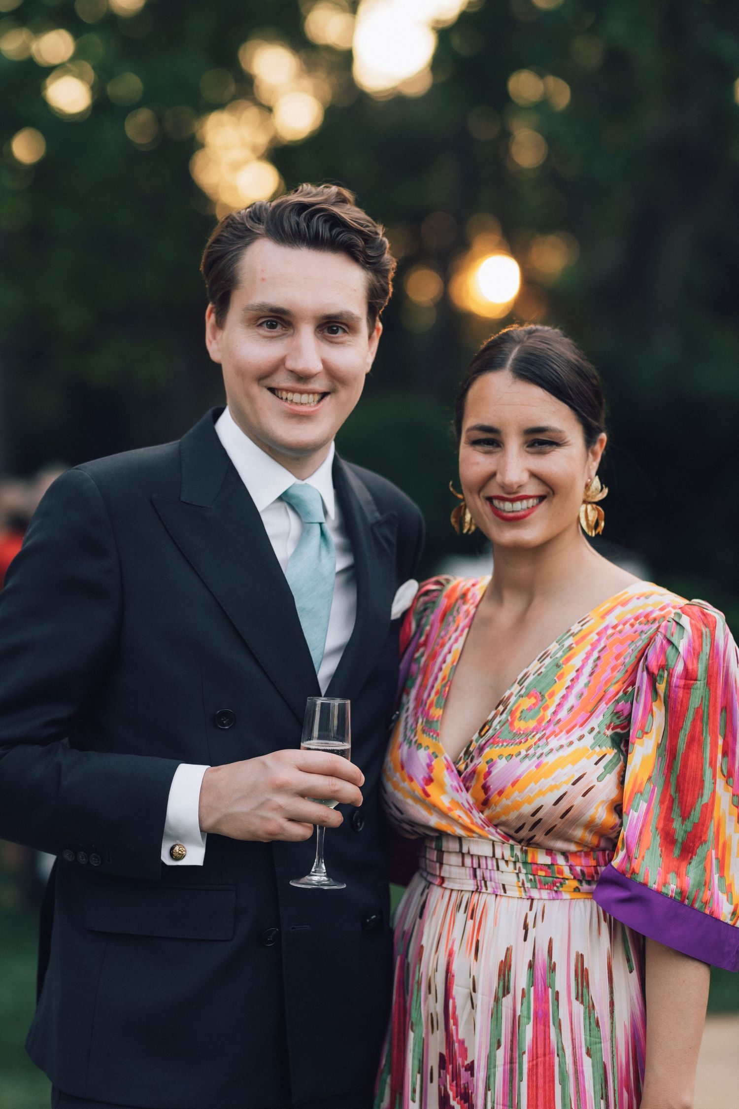 Couple souriant lors d'une soirée en plein air, homme en costume sombre avec champagne, femme en robe portefeuille imprimée colorée