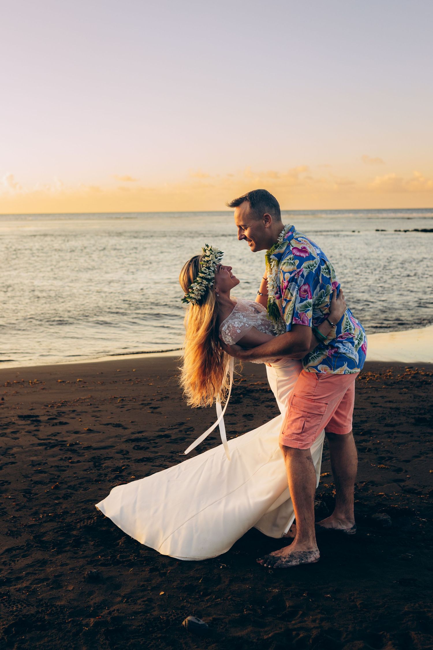 Couple en tenue de mariage dansant romantiquement sur une plage de sable noir au coucher du soleil à Hawaï.