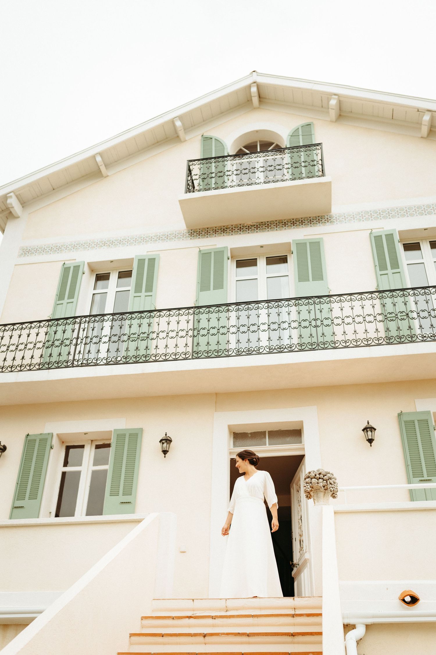 Mariée en robe blanche debout à l'entrée d'une élégante villa blanche avec volets verts et balcon en fer forgé en Provence