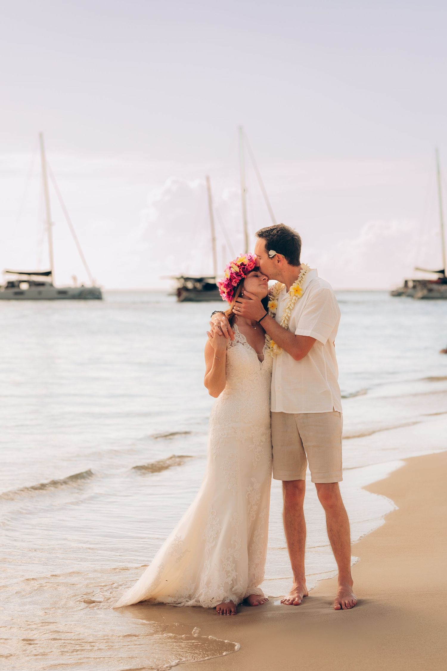 La mariée et le marié partagent un baiser romantique sur une plage tropicale avec des voiliers en arrière-plan.