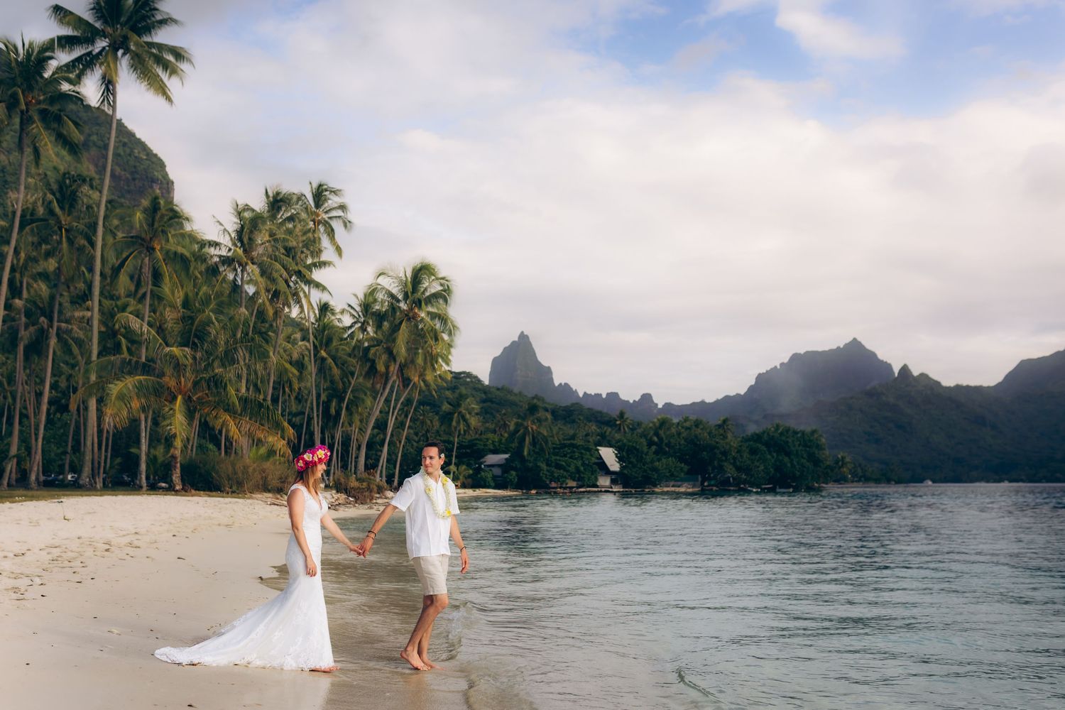 Mariée et marié se tenant la main marchant sur une plage de sable blanc avec des montagnes tropicales derrière.