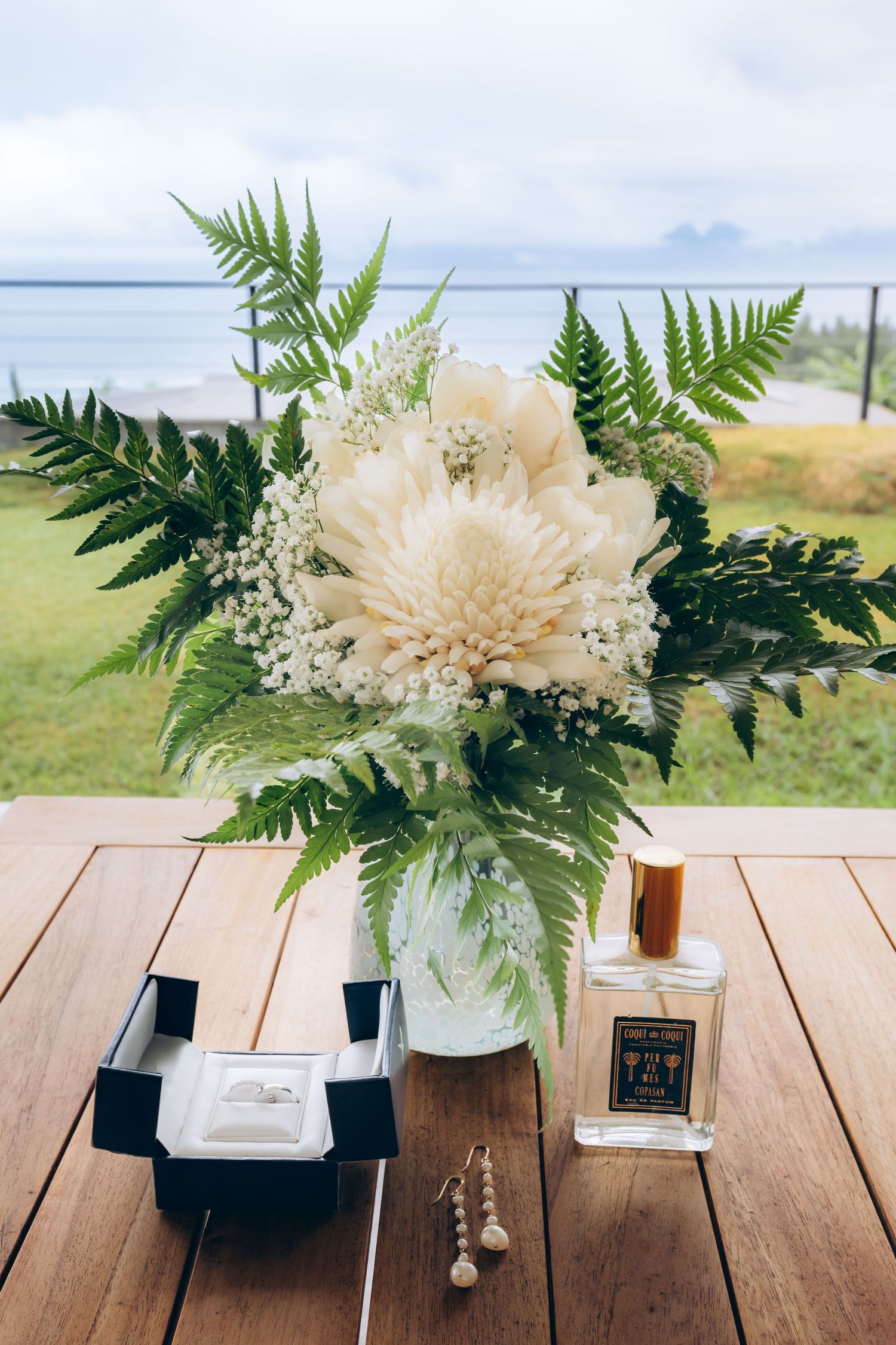 White dahlia wedding bouquet with ferns beside perfume bottle and jewelry box on wooden table outdoors.