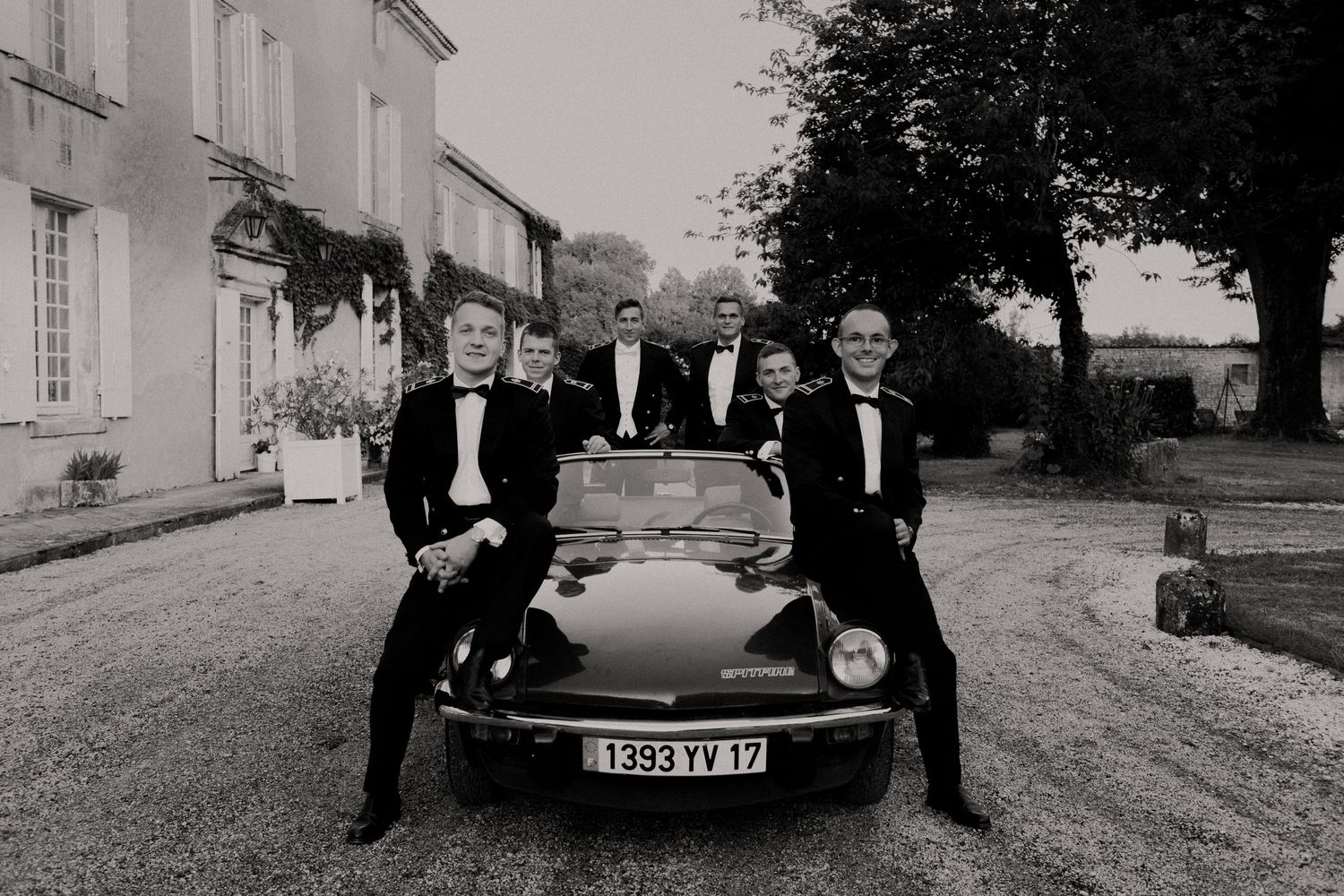 Cinq garçons d'honneur en smoking posant avec une voiture décapotable vintage à l'extérieur d'un château français en noir et blanc.