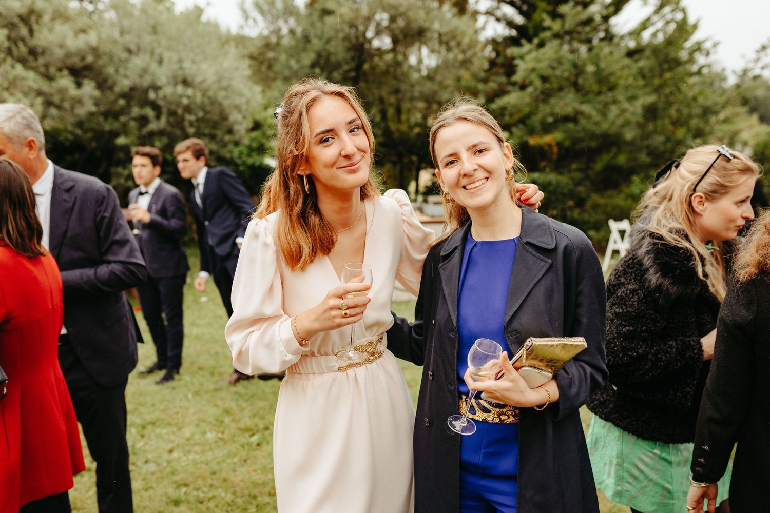 Deux femmes souriantes posant à l'extérieur lors d'une garden-party, l'une en robe blanche, l'autre en haut bleu avec un blazer foncé.