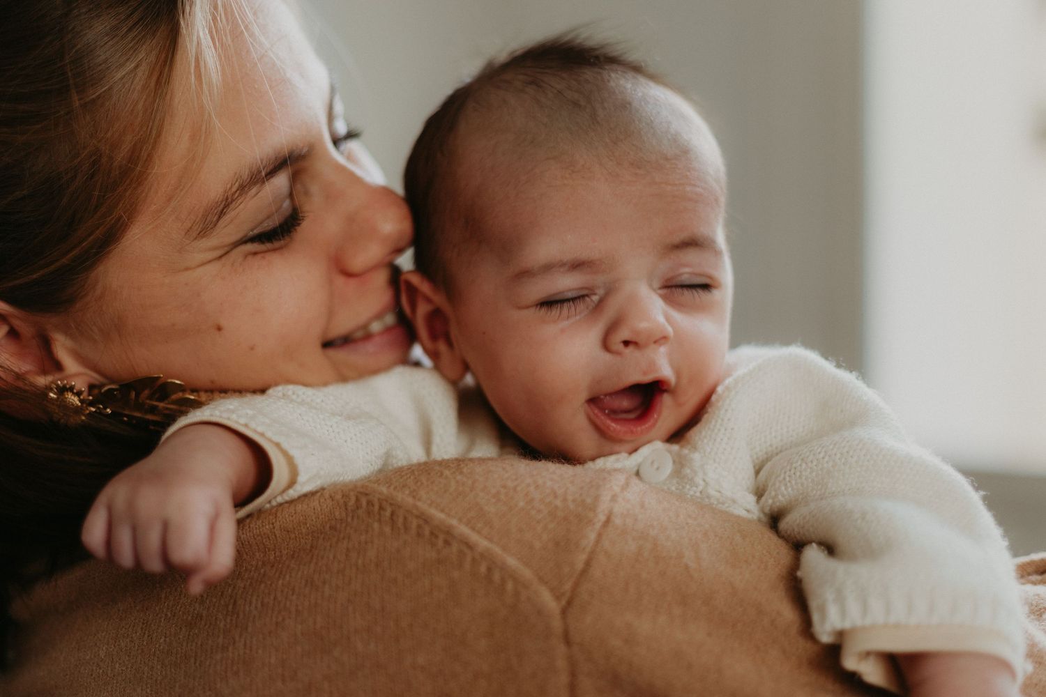 Une mère souriante tient son nouveau-né heureux et riant vêtu d'un pull en tricot blanc.