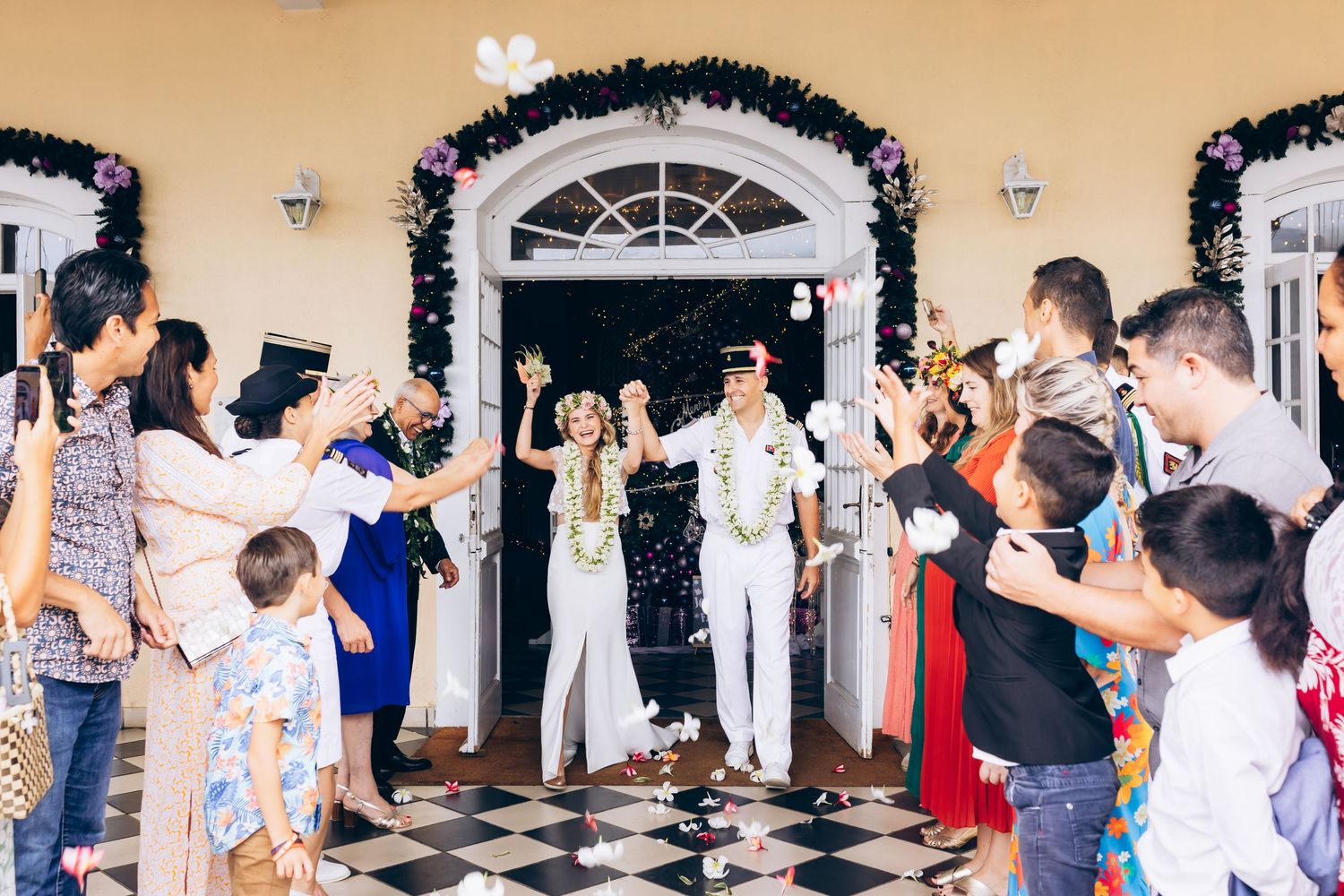 Un couple de mariés traverse des invités acclamant en jetant des confettis à l'extérieur d'un lieu magnifiquement décoré.