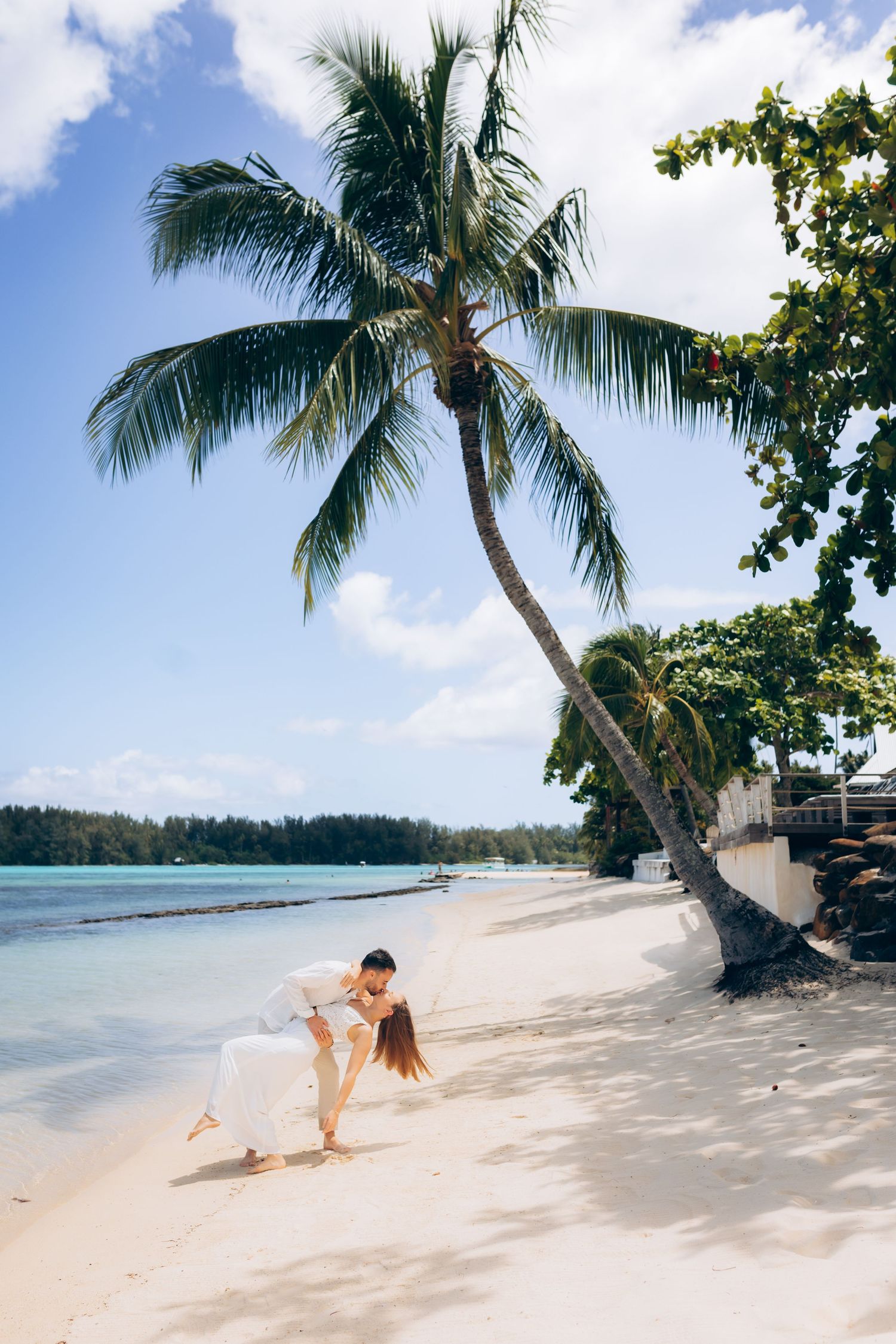 Marié plongeant la mariée en robe blanche sur une plage tropicale avec un palmier penché.
