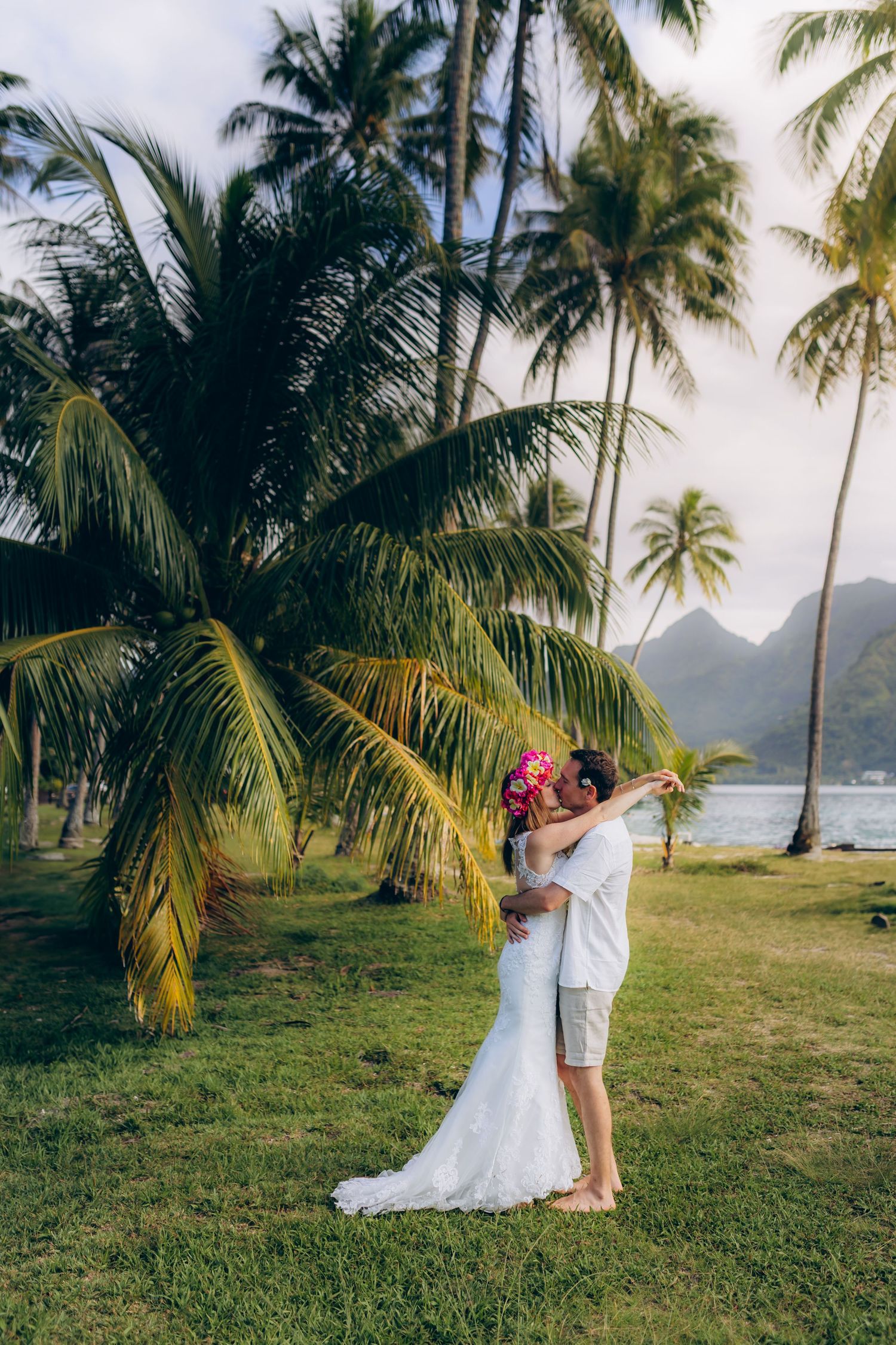 Couple de marié s'embrassant sous un grand palmier avec des montagnes tropicales et un océan en arrière-plan.