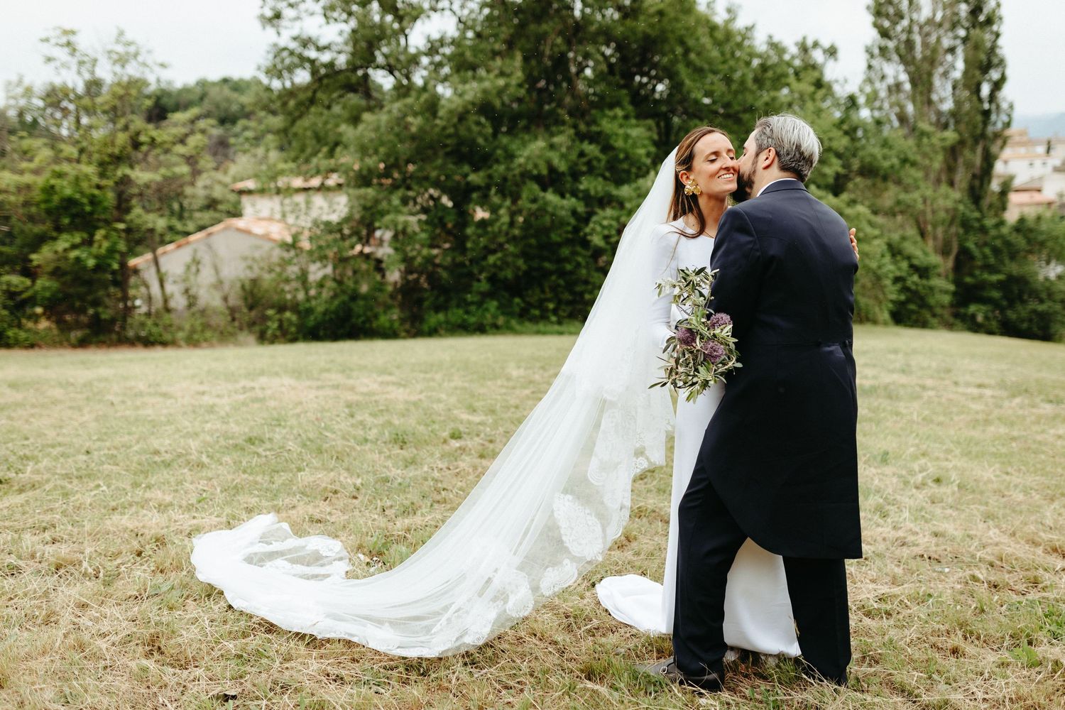 La mariée et le marié partagent un baiser romantique à l'extérieur sur une colline herbeuse, avec un long voile blanc en dentelle