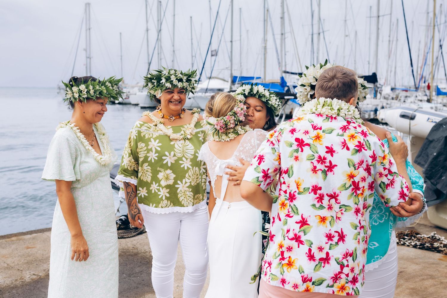 Les invités de mariage en tenue florale se mêlent près d'une marina avec des voiliers lors d'une célébration tropicale.