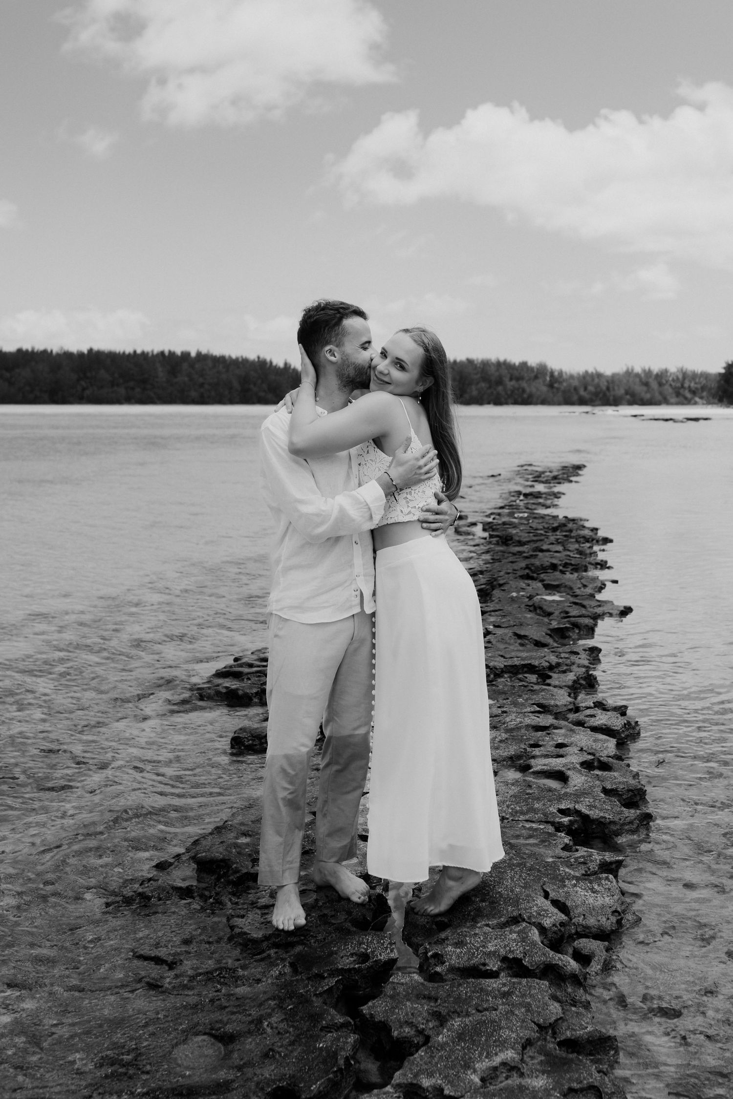 Photo en noir et blanc d'un couple s'embrassant sur un chemin rocheux entouré d'eau de l'océan.