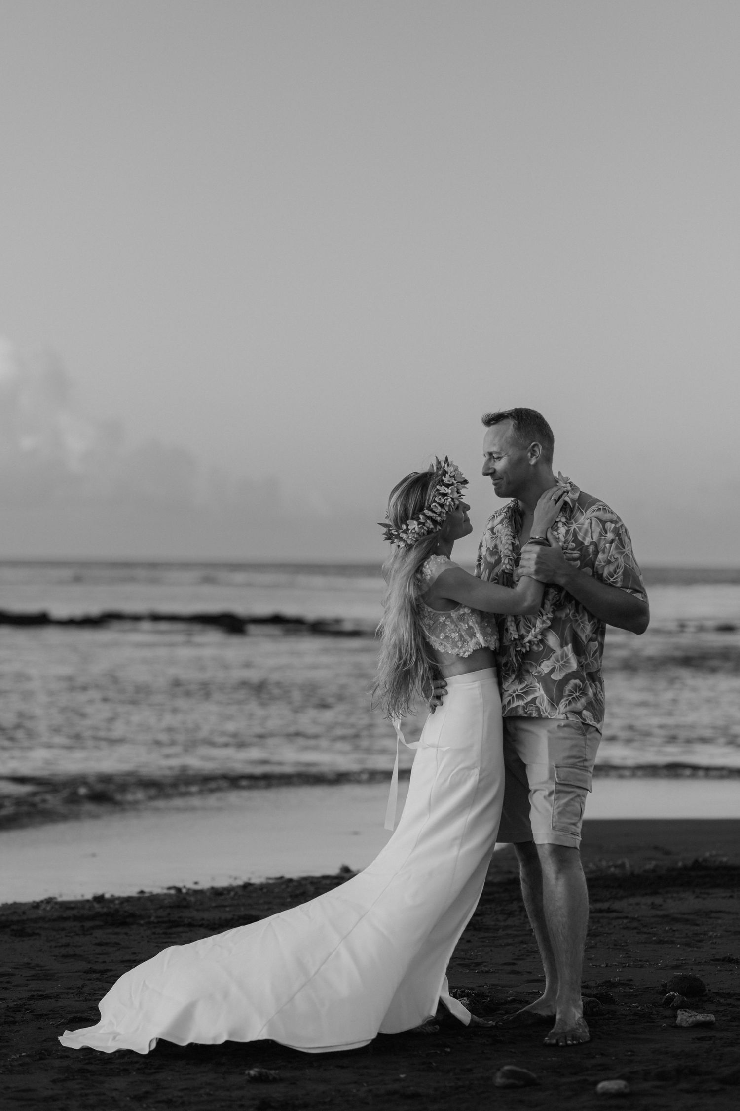 Photo en noir et blanc d'une mariée et d'un marié s'embrassant sur une plage au coucher du soleil.