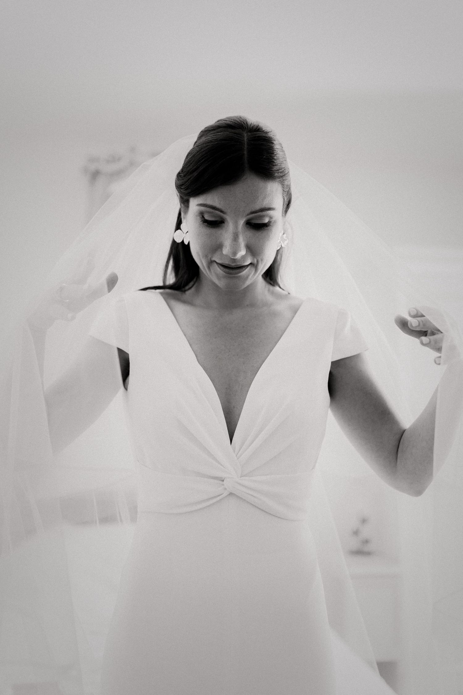 Black and white portrait of smiling bride in deep V-neck gown holding wedding veil.